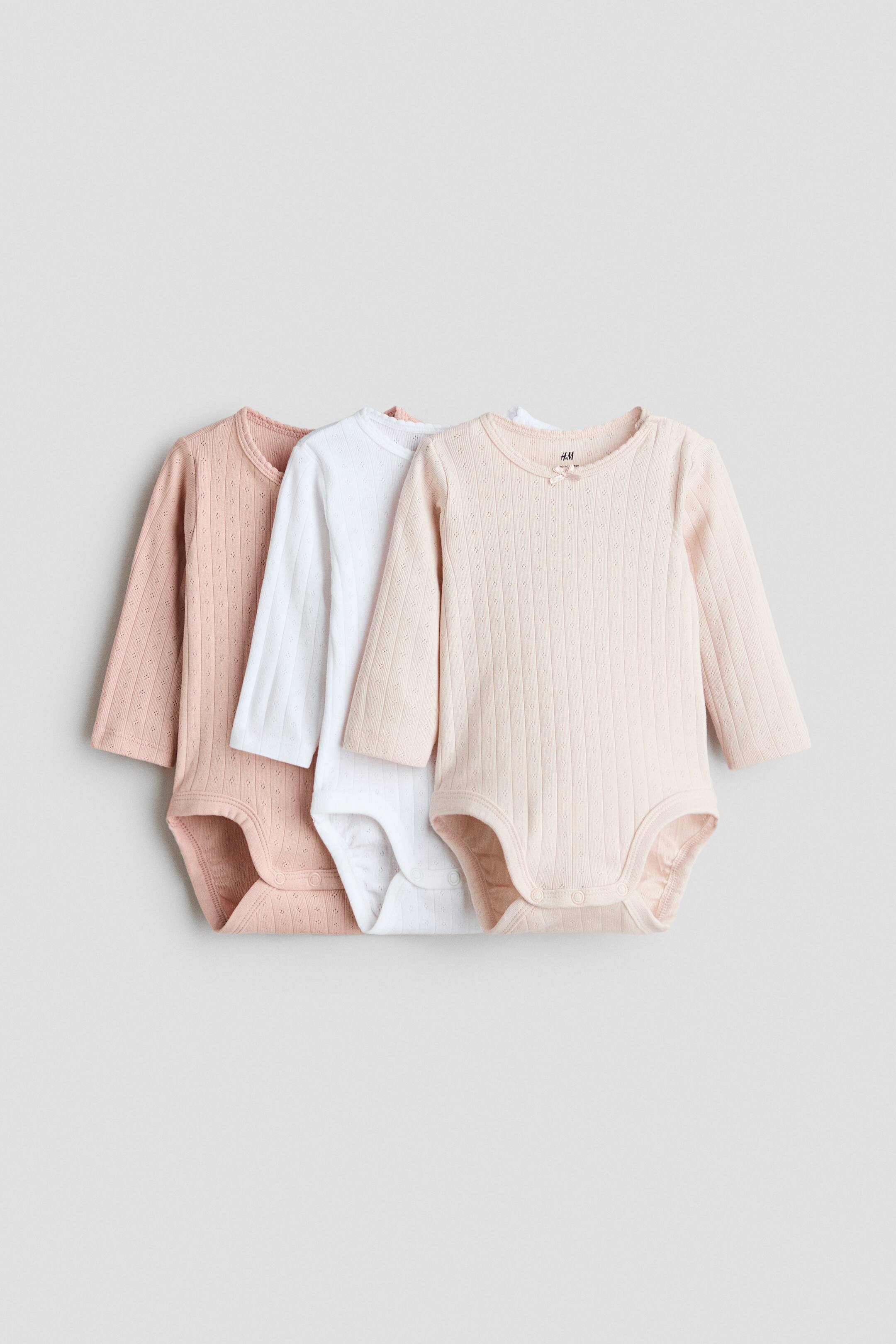 Xem ảnh lớn hơn: Ba áo liền quần tay dài cho bé bằng chất vải pointelle jersey được bố trí chồng lớp, hướng mặt trước, với các màu hồng phấn, trắng và hồng đào nhạt. Mỗi áo đều có cổ tròn viền lượn sóng, trang trí một nơ nhỏ ở giữa, tay dài và cúc bấm ở phần đáy quần.