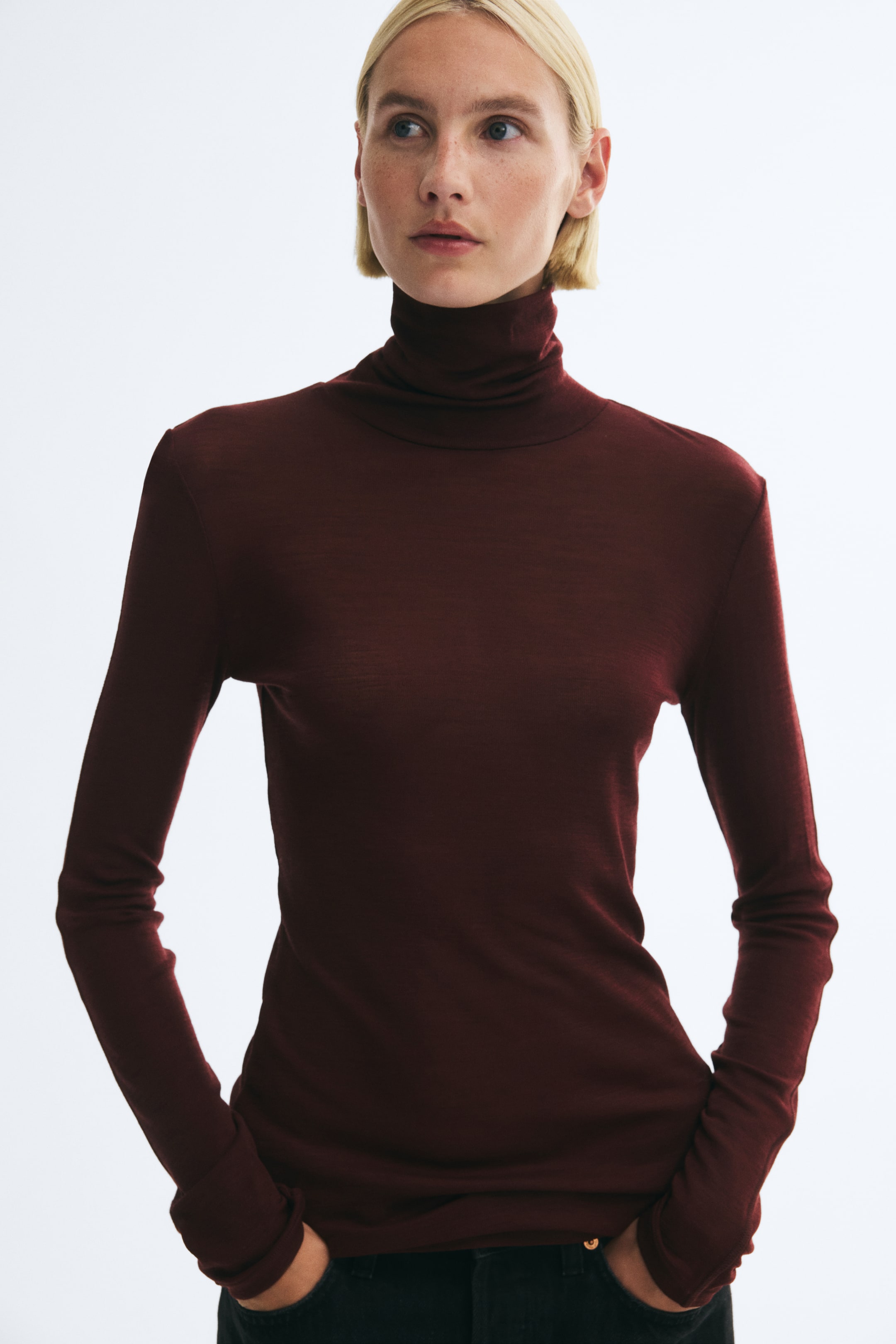 Grotere afbeelding bekijken: Het model draagt een nauwsluitende, donker bordeauxrode coltrui van fijne brei. Het topje met lange mouwen is gestyled met een donkere broek, terwijl het model haar handen in de zakken heeft geplaatst.