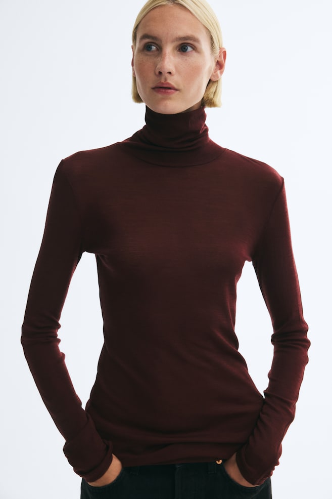 A fitted, dark burgundy roll-neck top in a fine knit is worn by the model. The long-sleeved top is styled with dark trousers, with the modelâs hands placed in the pockets. - 1