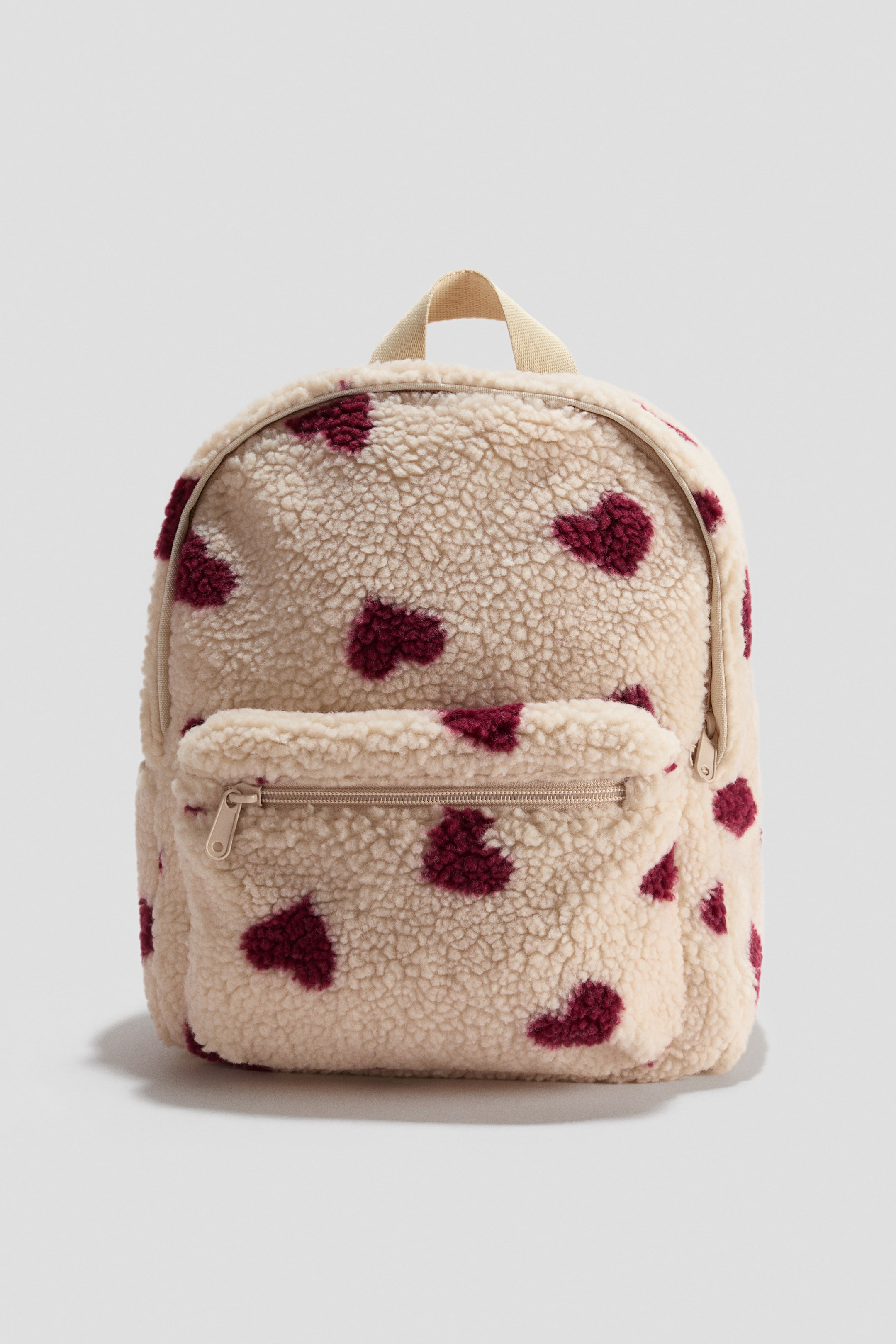 View larger image: A cream-colored sherpa backpack displays scattered burgundy heart patterns on the front. It features a zippered main compartment, a front zippered pocket, and a cream fabric loop handle at the top.