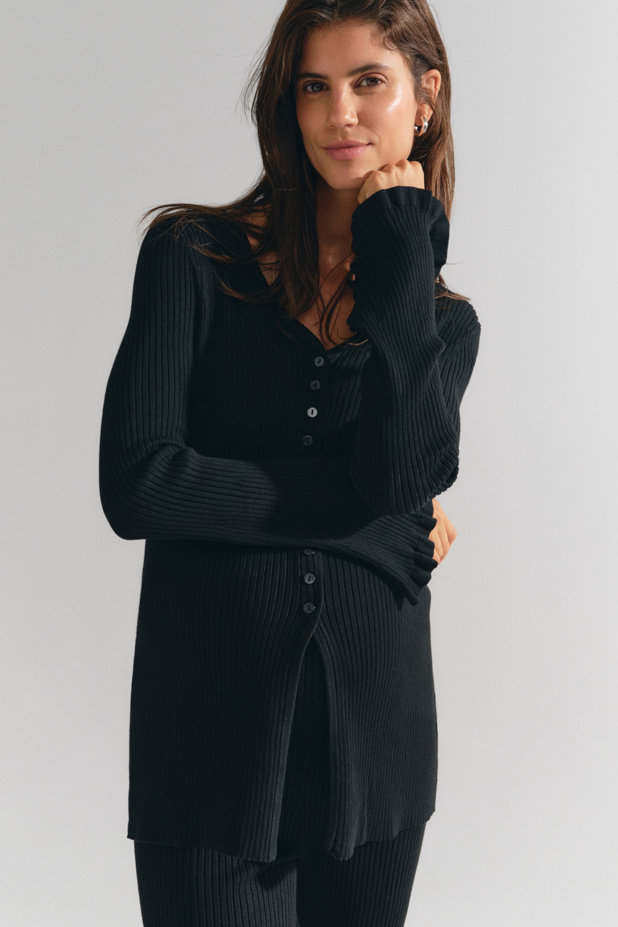 View larger image: A long, black, fitted rib-knit cardigan featuring a V-shaped neckline, front button closures, and long sleeves with frill-trimmed cuffs is worn by the model. The model is pairing the cardigan with matching black rib-knit trousers, posing with one arm crossed over the torso and one hand resting under the chin.
