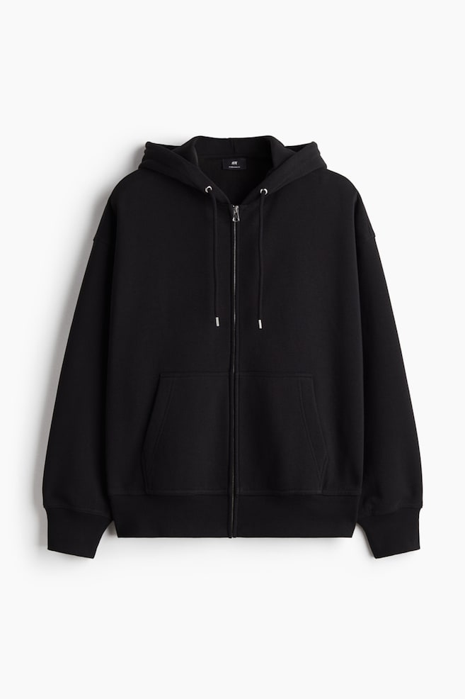 A black zip-through hoodie is presenting front, featuring an oversized fit with dropped shoulders. It is designed with a full zipper closure, a drawstring hood, long sleeves, front pockets, ribbed cuffs, and a ribbed hem. - 1