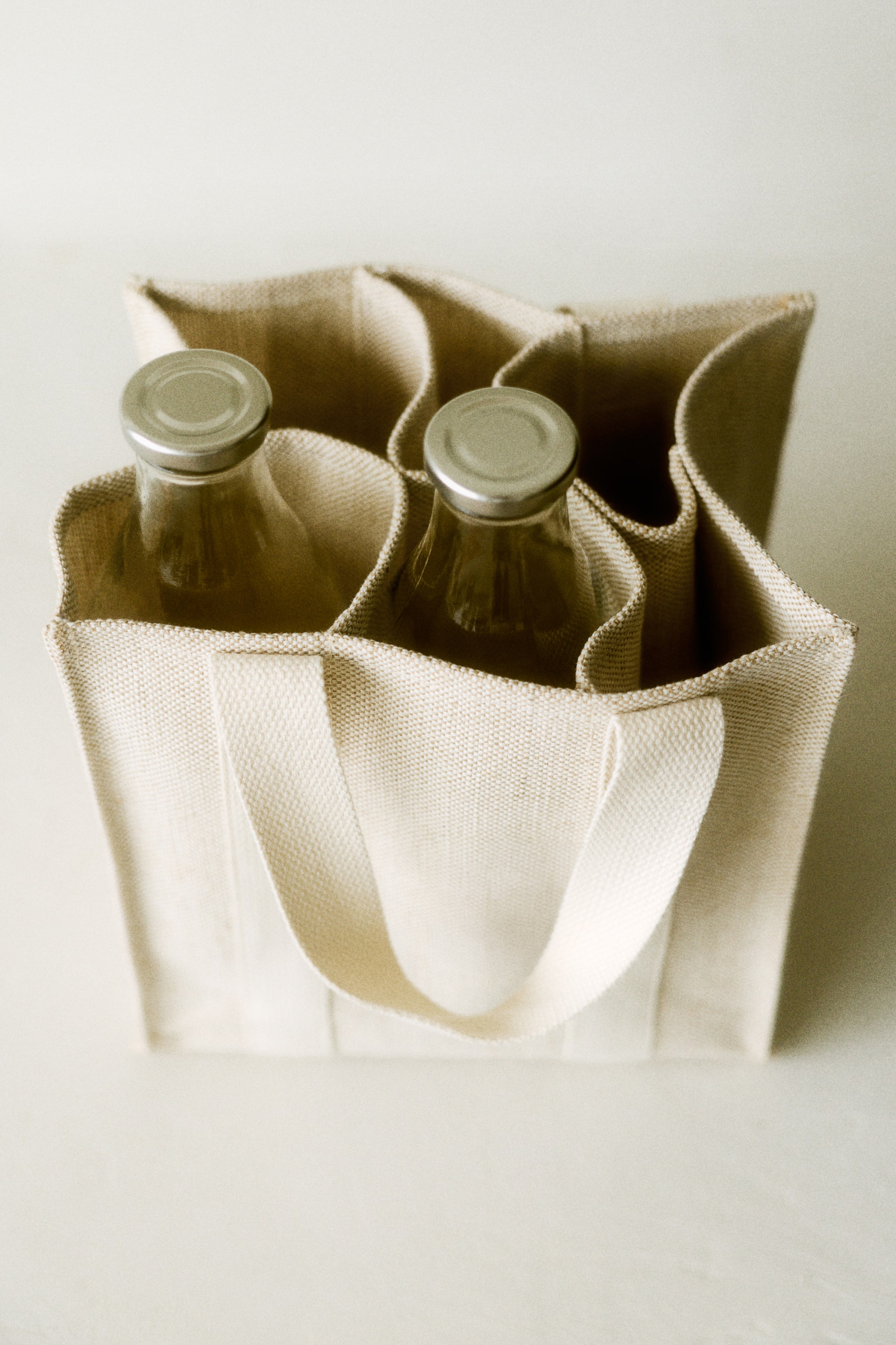 Ver imagen más grande: Una bolsa tote de lona de color natural está ligeramente inclinada hacia el espectador, mostrando sus cuatro compartimentos internos y asas de tela tejida, con dos botellas de vidrio transparente, tapadas con plata, colocadas dentro de dos de los compartimentos.
