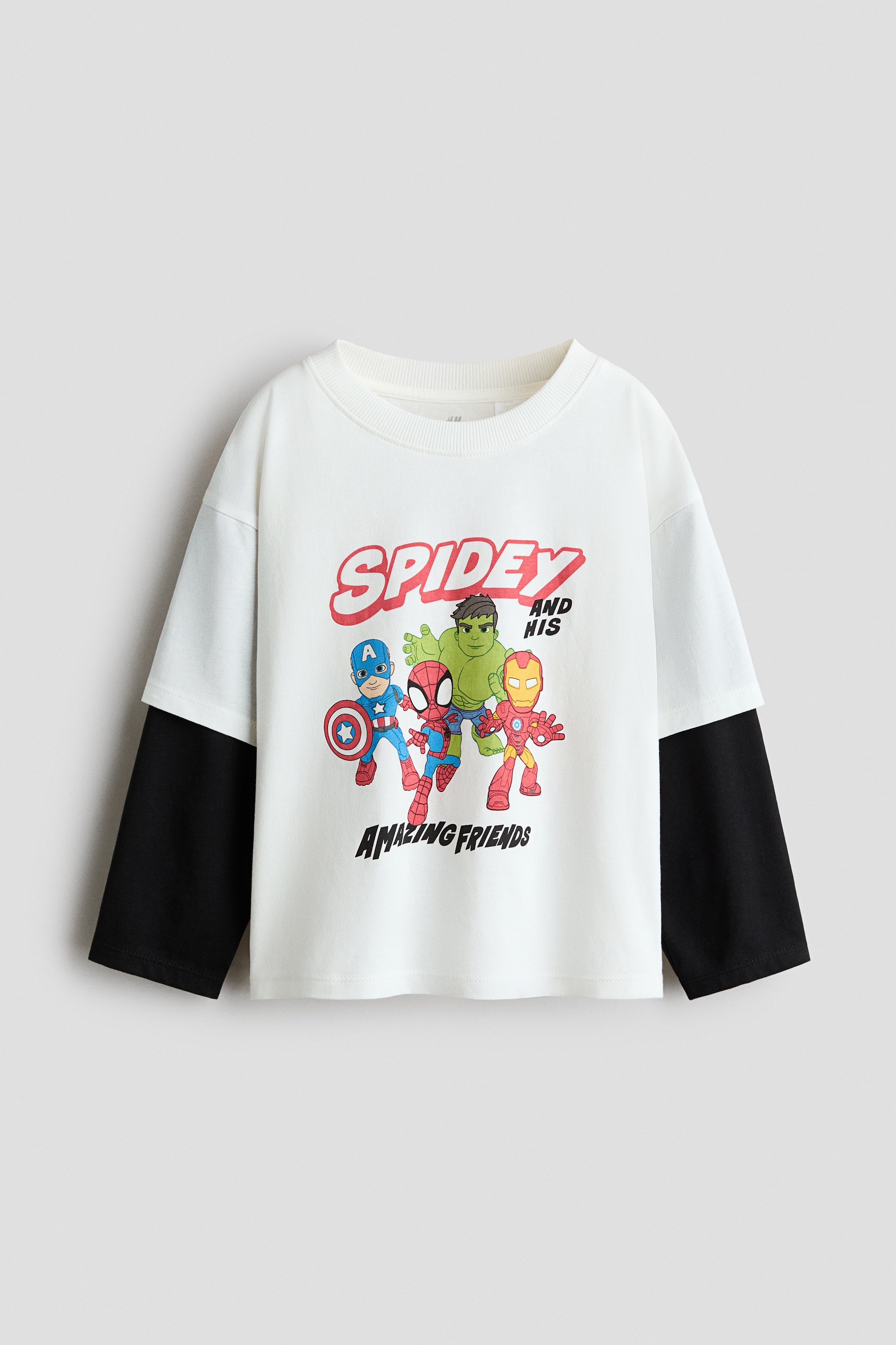 A white graphic T-shirt with a cartoon print is worn by the model, who is holding the hem. The T-shirt features dropped shoulders, a ribbed crew neck, and a layered design with short sleeves over sewn-in long black sleeves.