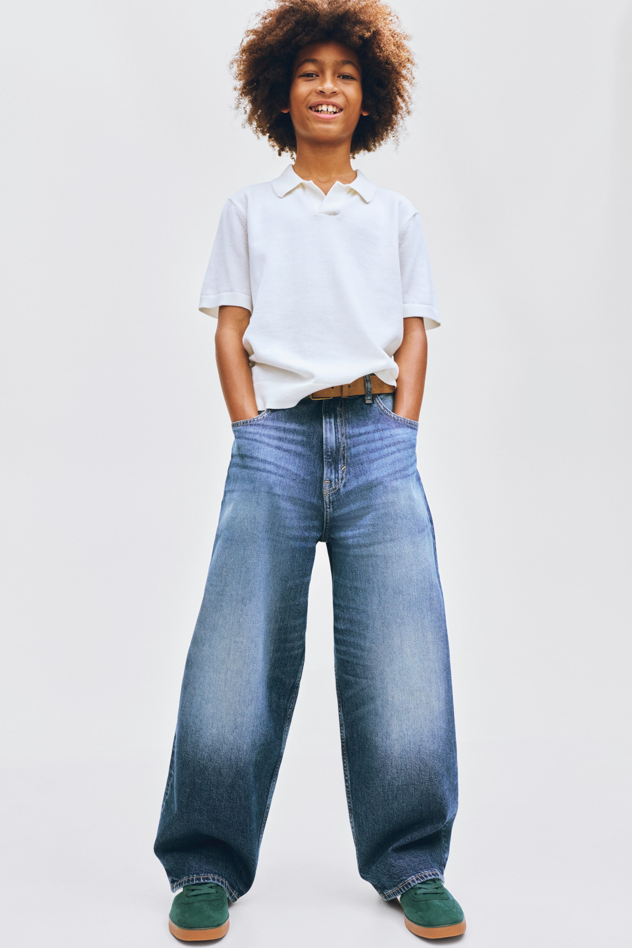 Voir une image plus grande: Un polo blanc à manches courtes, avec un col classique et une patte de boutonnage, est porté non rentré sur un jean bleu à jambes larges. Le jean présente un délavage clair qui passe à une teinte plus foncée aux ourlets et est maintenu par une ceinture beige.