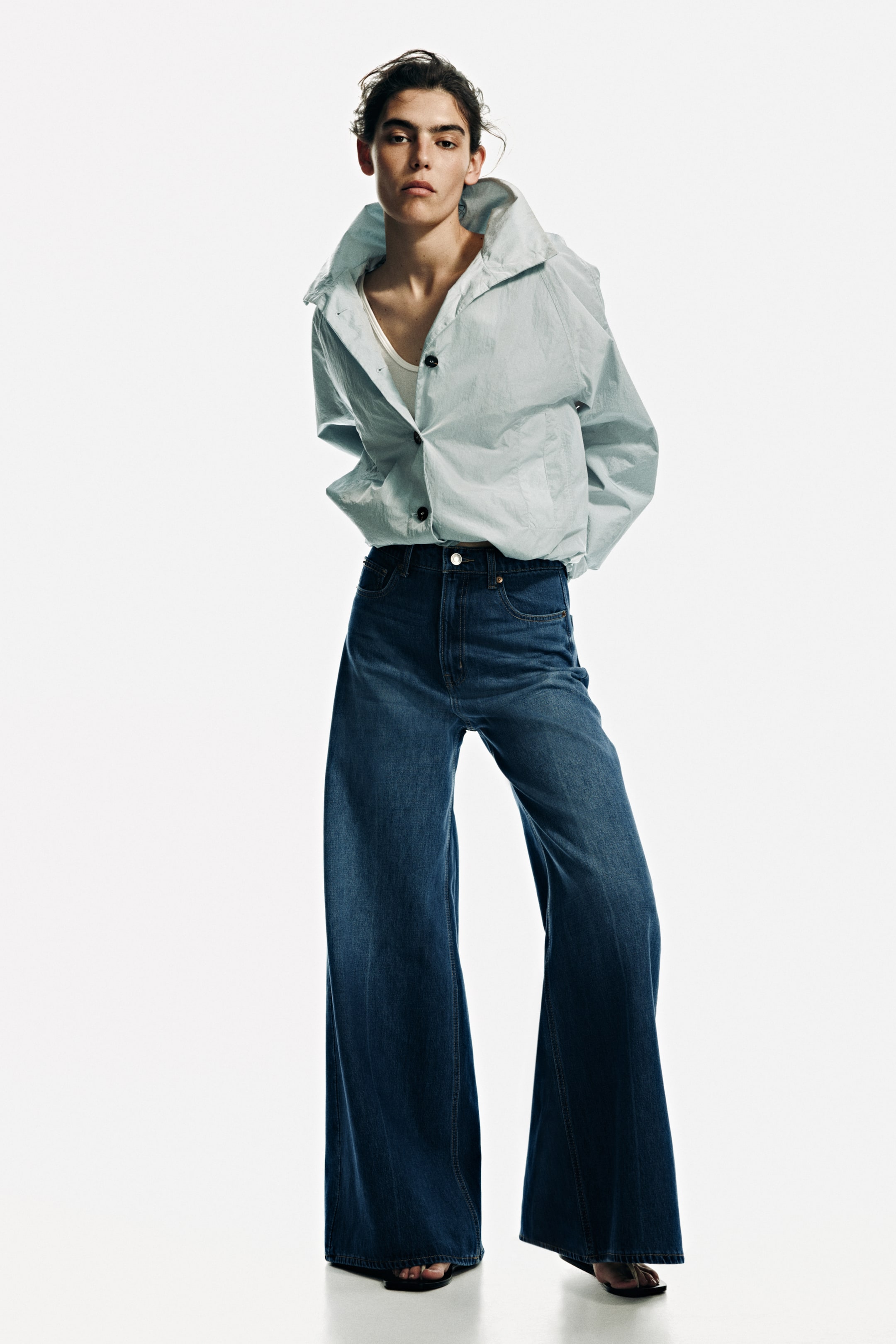 View larger image: The model is showcasing 5-pocket jeans, characterized by a dark blue wash, high waist, and a regular fit around the hip area. These jeans boast a wide leg design with an extra-wide cut extending from the thigh to the hem. They feature a visible zip fly and button closure, and their regular length gracefully touches the top of the model's foot. Complementing the look, the model is adorned in a light blue jacket and sandals.