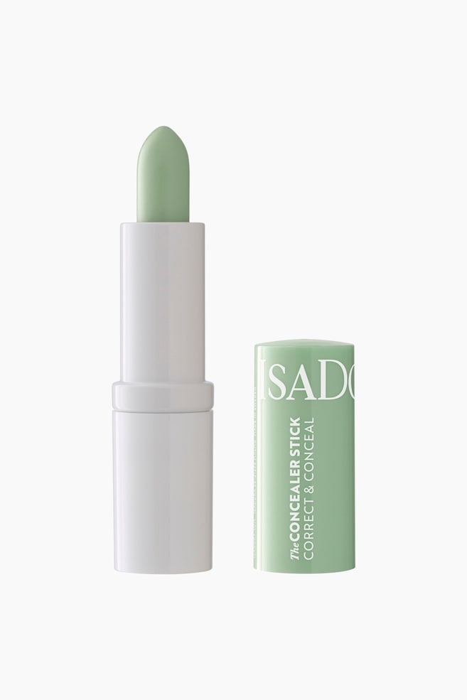 En grønn concealer-stift står oppreist, der det spisse, lysgrønne produktet er trukket ut fra en hvit base. Til høyre for den står det matchende lysgrønne lokket, med teksten "ISADORA" og "THE CONCEALER STICK CORRECT & CONCEAL", også oppreist. - 1