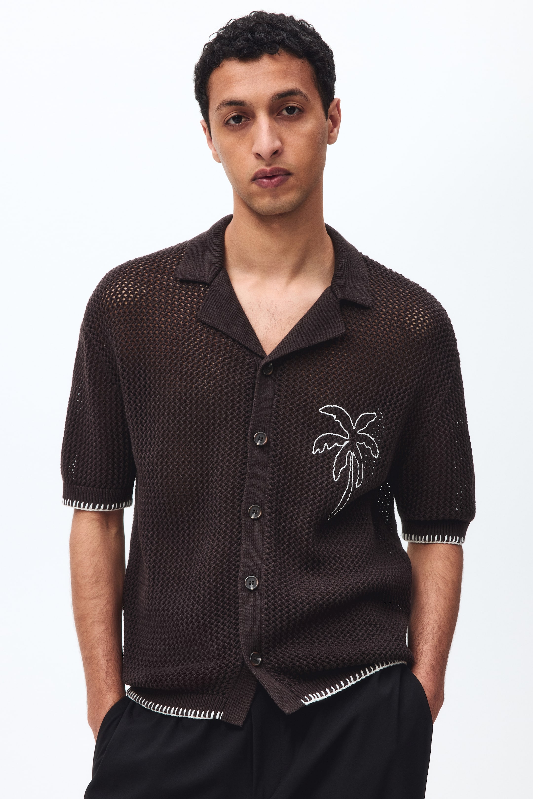 View larger image: The model is wearing a brown, short-sleeved, hole-knit shirt with a resort collar and a relaxed fit, standing with his hands in his pockets. The shirt showcases a white embroidered palm tree on the left chest, buttons down the front, and features decorative white blanket stitching at the cuffs and straight-cut hem.