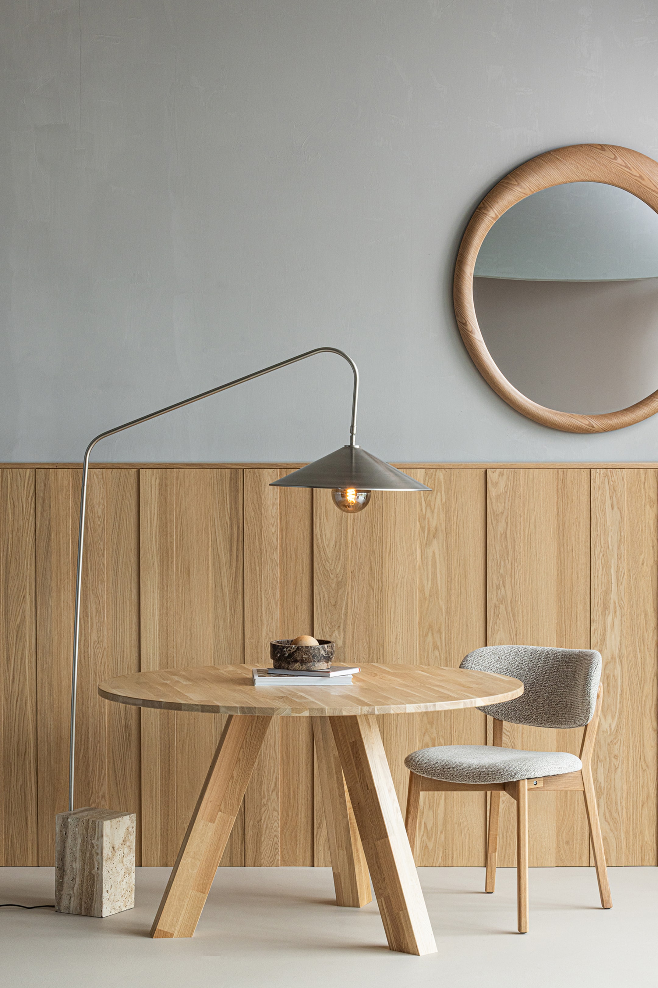 Grotere afbeelding bekijken: De ronde houten eettafel toont een glad, lichtgetint houten oppervlak met zichtbare nerf. Hij wordt ondersteund door vier robuuste, schuin geplaatste houten poten, en op het blad staat een donkere, patroonrijke schaal met lichte objecten en een stapel witte boeken.