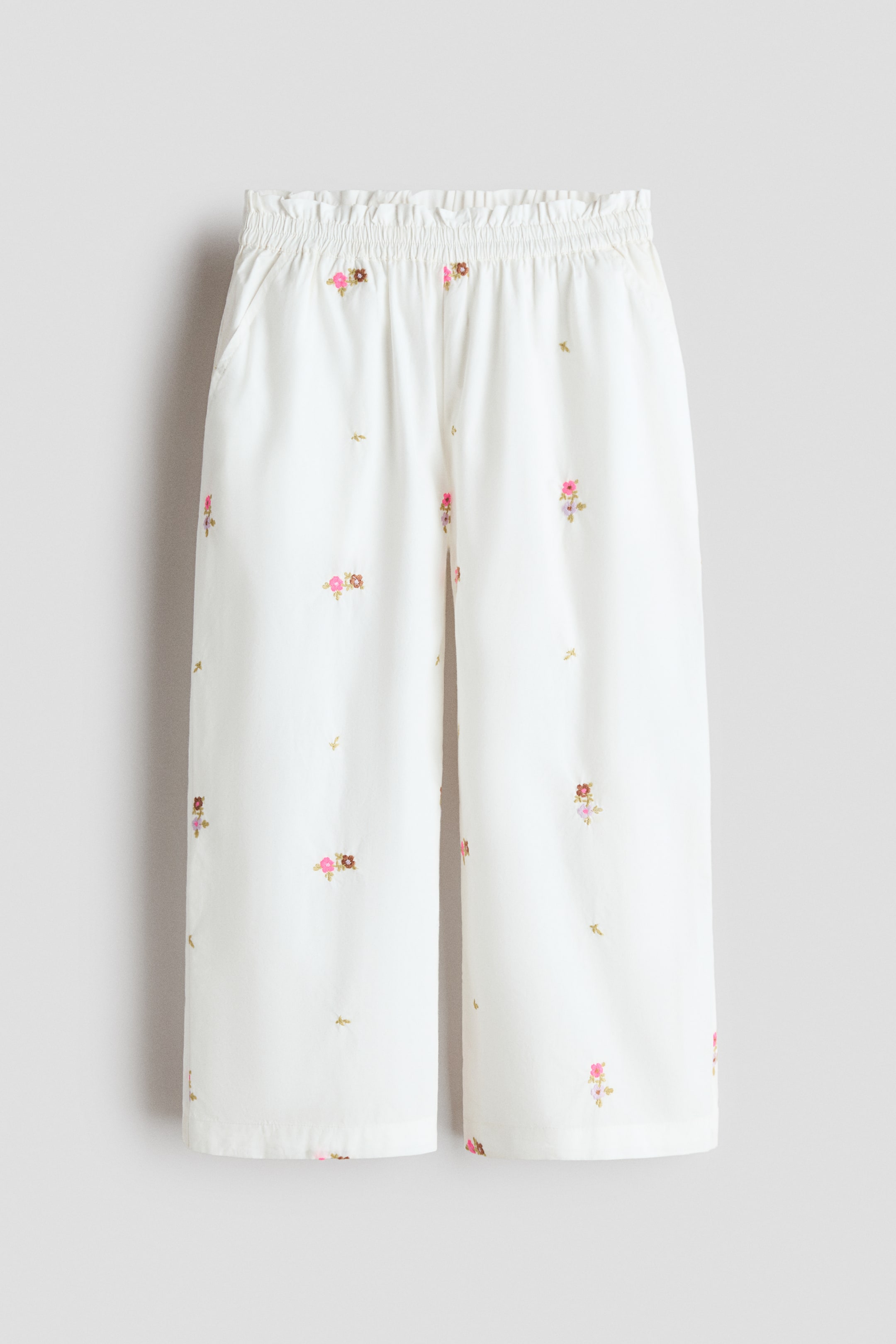 Visa större bild: Vita vida byxor med breda ben har en rynkad elastisk midja, sidfickor och blomsterbroderier i rosa och bruna nyanser med små gyllene motiv, presenterade framifrån.