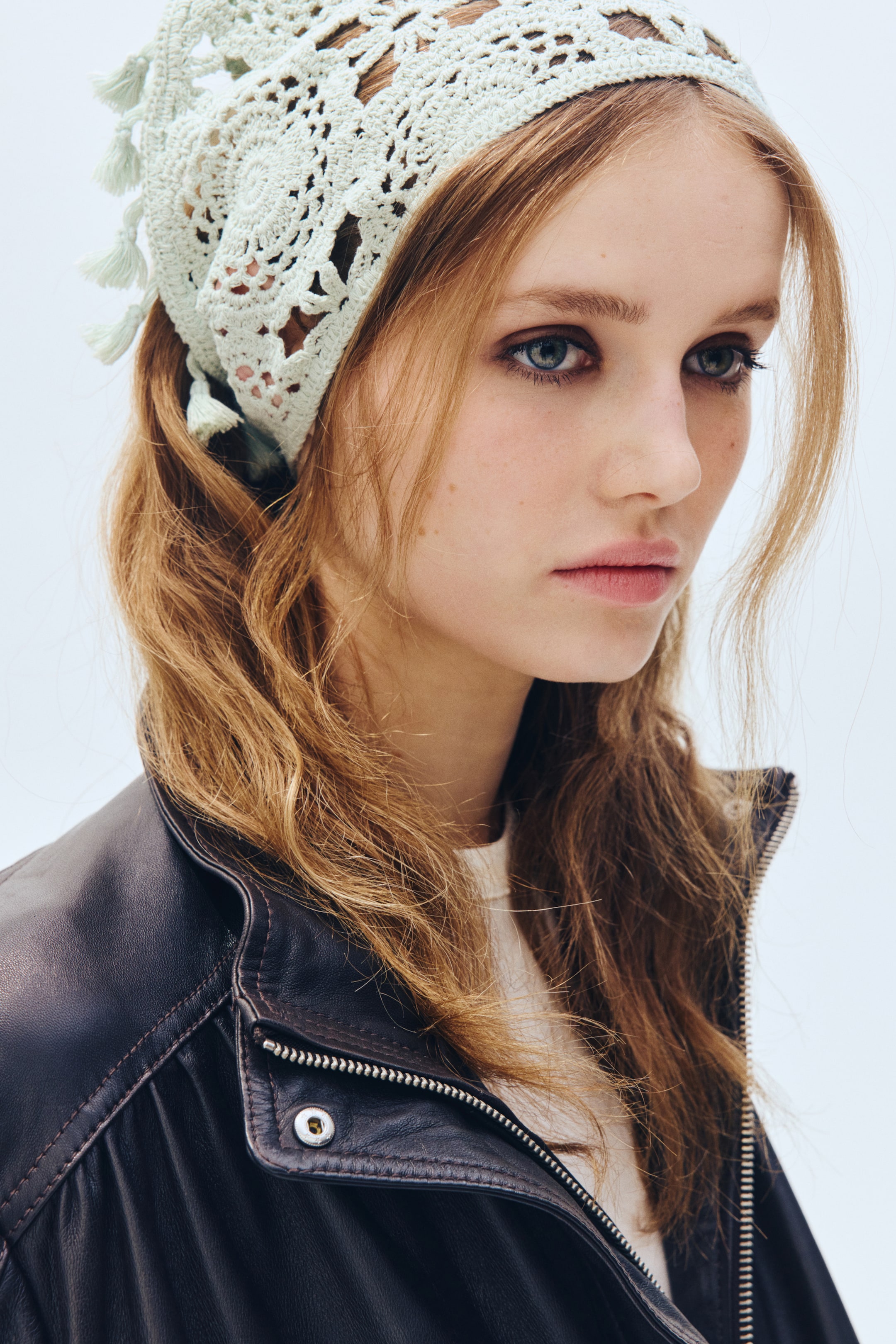 View larger image: The light green crochet headscarf gracefully drapes over the top of the head, extending from the forehead to the back. Showcasing an intricate open-knit pattern, it is adorned with small tassels along its visible edges.