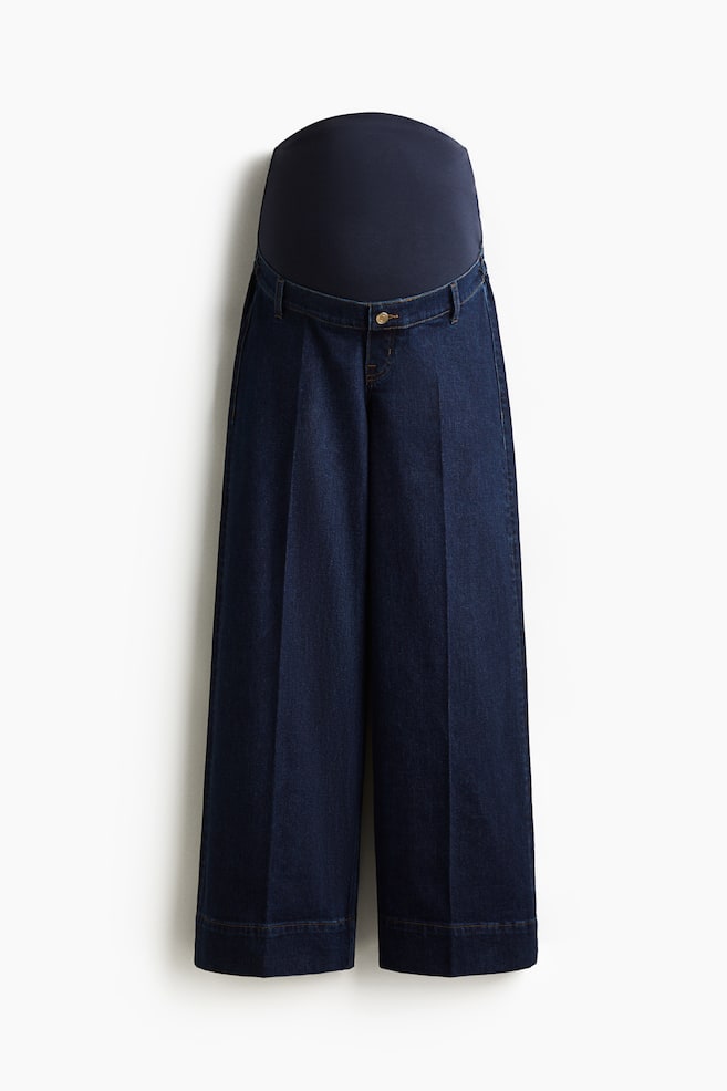A pair of dark blue maternity jeans are presenting from the front, featuring a wide-leg cut with visible creases down the front. They are showing a wide, dark blue jersey panel at the waist, a denim waistband with belt loops, and a single button closure. - 1