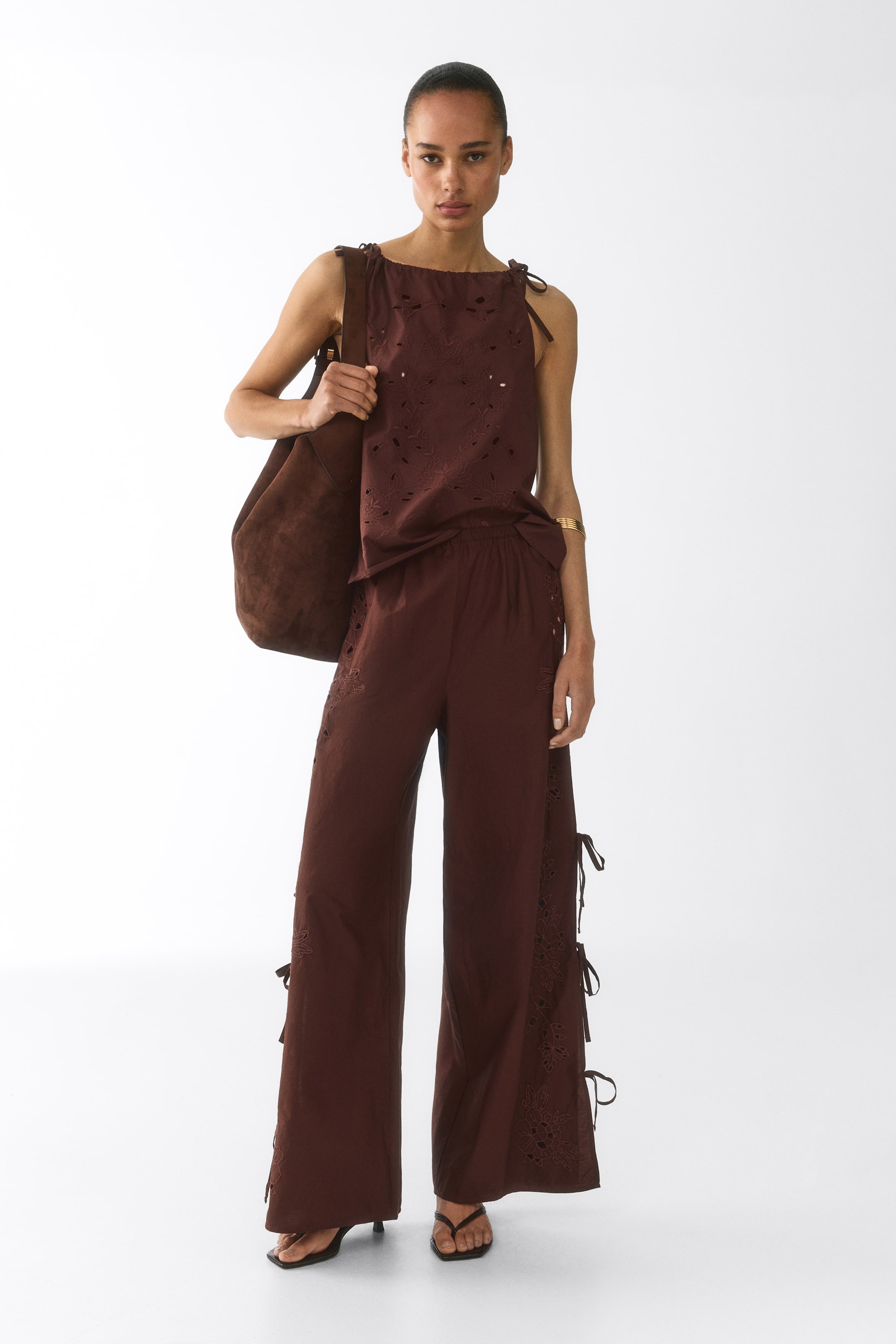 View larger image: A dark brown sleeveless top and matching wide-leg pants are showcased in a front view. The top highlights intricate eyelet embroidery and tie details on the shoulders, while the pants are crafted with an elasticated waistband, eyelet embroidery, and side tie details along the outer seams.