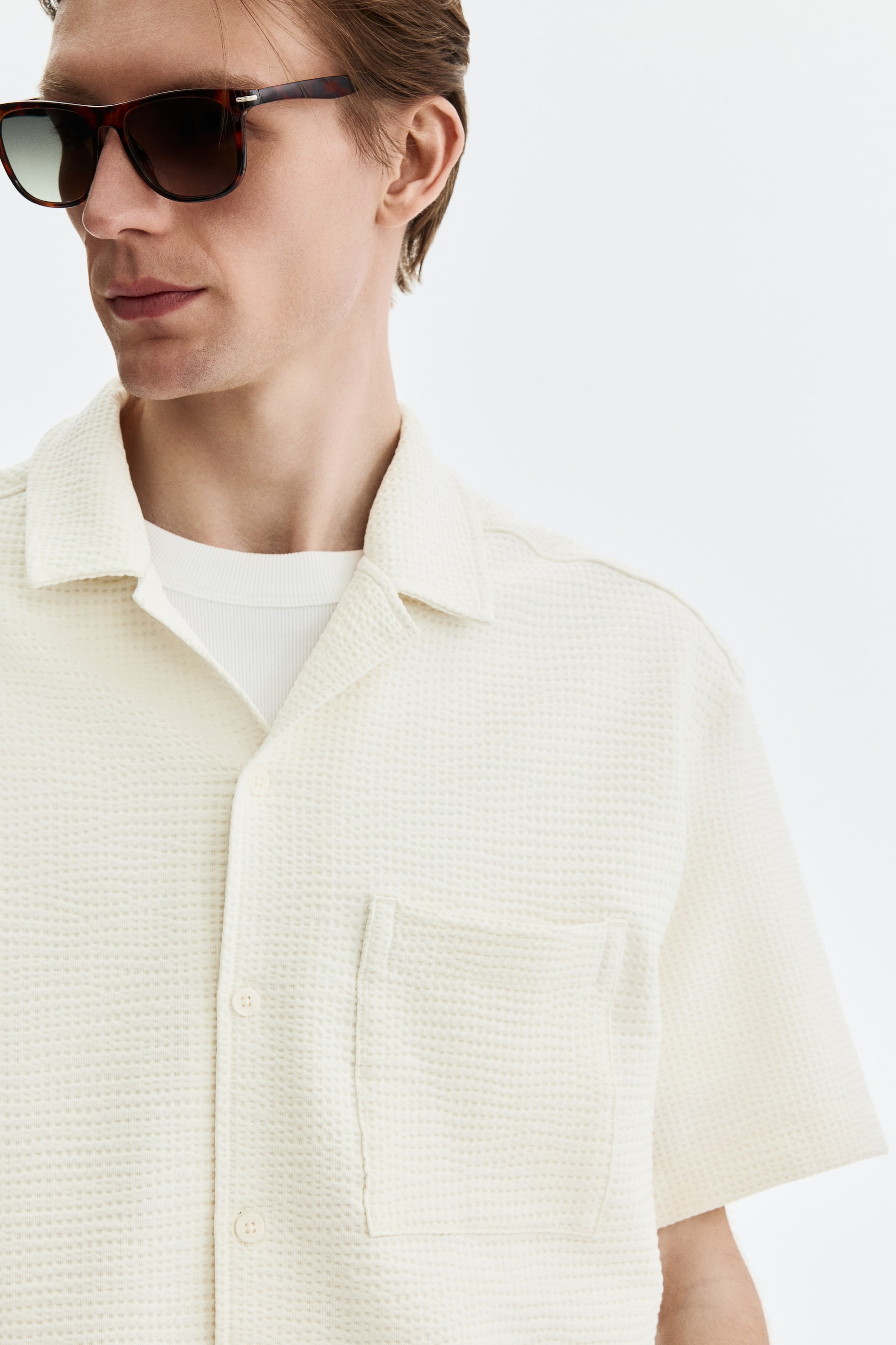View larger image: A cream waffle-knit short-sleeve shirt is being worn, shown from a slight front-right angle. It features an open camp collar, a buttoned placket, and a left chest patch pocket, with a white ribbed undershirt visible underneath.