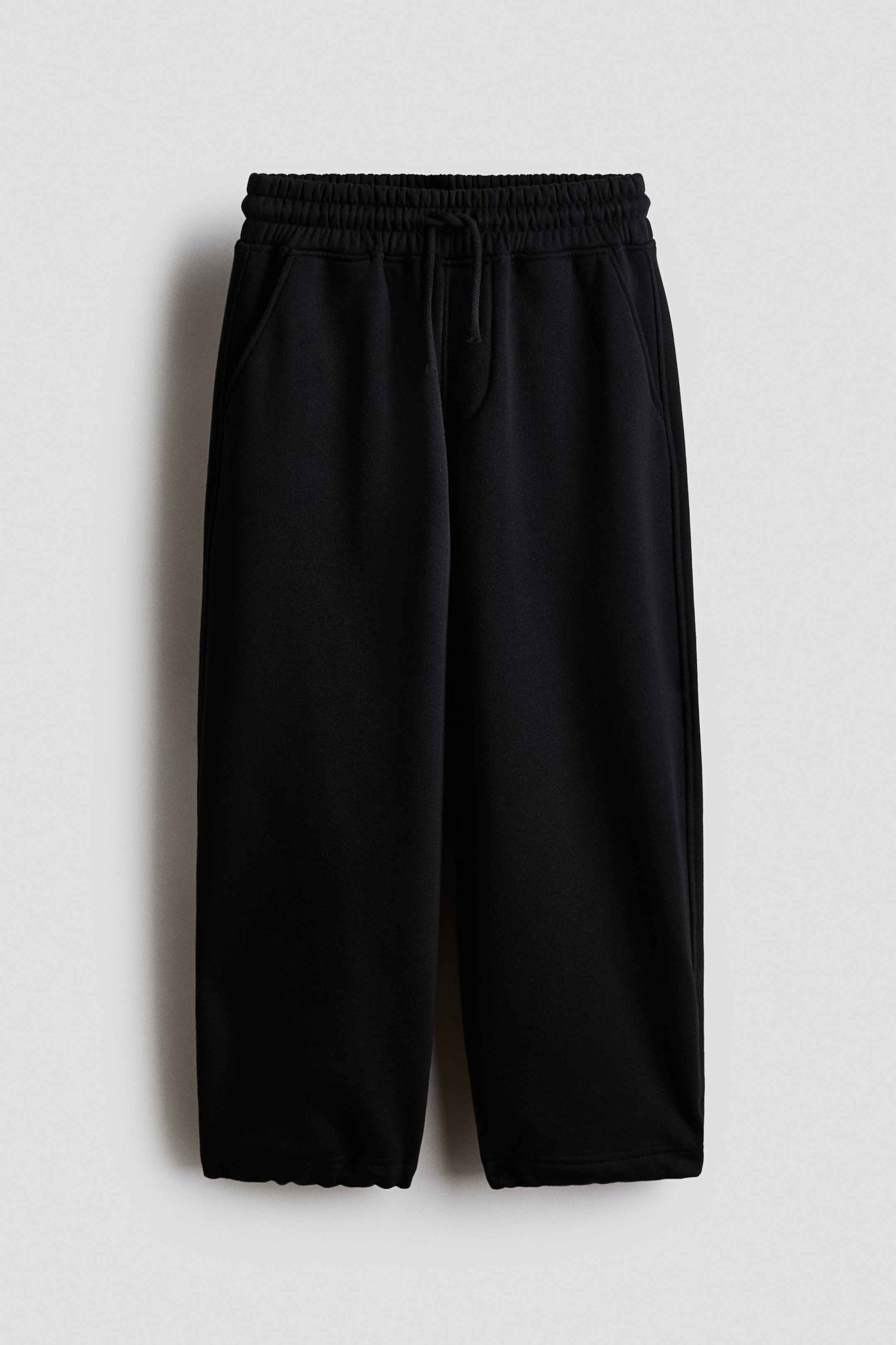 Größeres Bild ansehen: Schwarze Jogginghosen aus Sweatshirt-Stoff werden von vorne präsentiert und verfügen über einen elastischen Bund mit Kordelzug und einen Fake-Schlitz. Sie zeigen schräge Seitentaschen und weite, ballonförmige Beine mit elastischen Säumen.