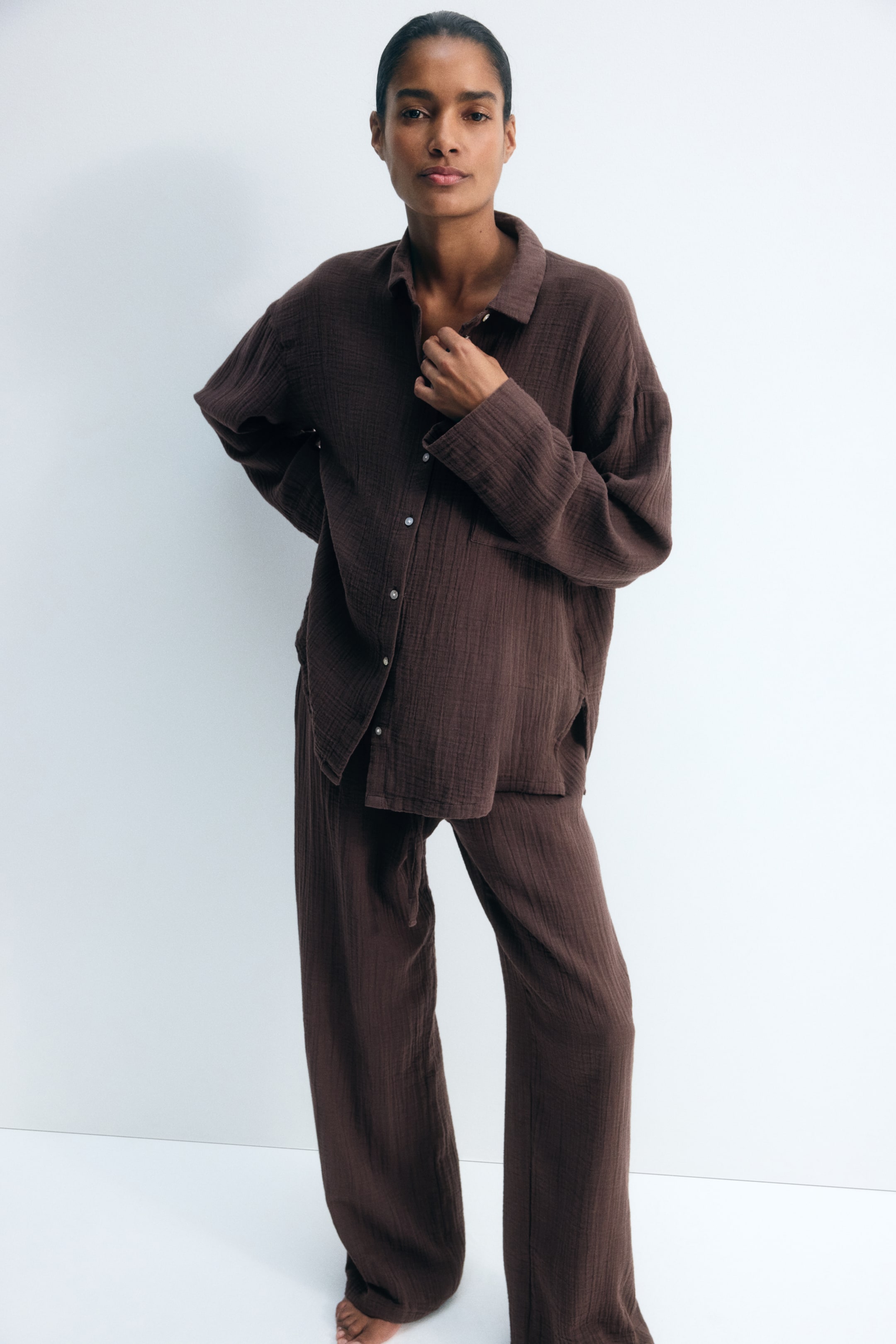 Agrandir l'image: Un ensemble de loungewear à texture froissée, de couleur marron foncé, est porté par le mannequin, qui ajuste le col de la chemise à manches longues d’une main. L’ensemble se compose d’une chemise oversize boutonnée avec une poche poitrine et d’un pantalon large assorti à coupe ample.