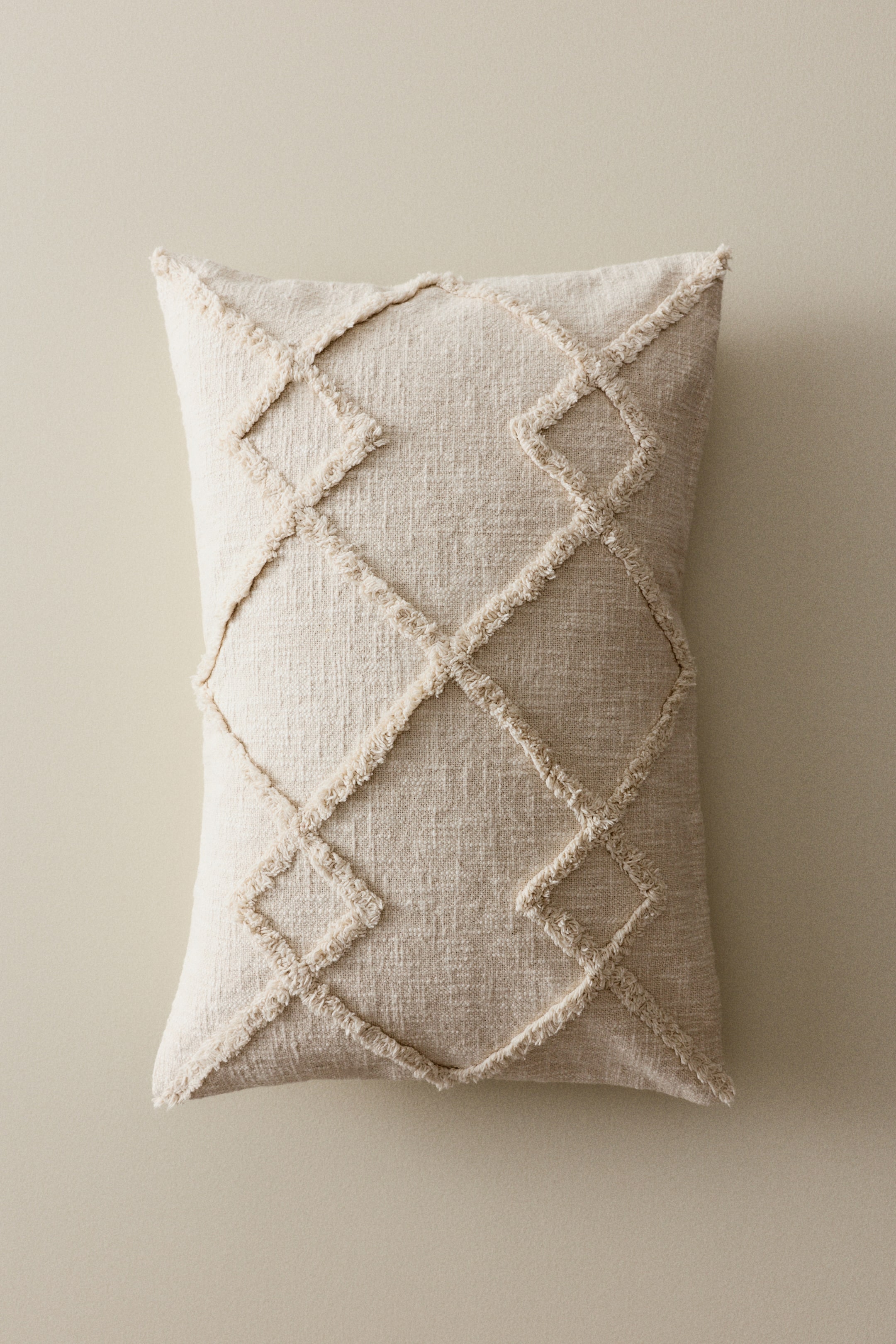 拡大画像を表示: ライトベージュの長方形の枕は、織り目のある表面に、立体的でほつれた幾何学的なダイヤモンド模様が施されています。