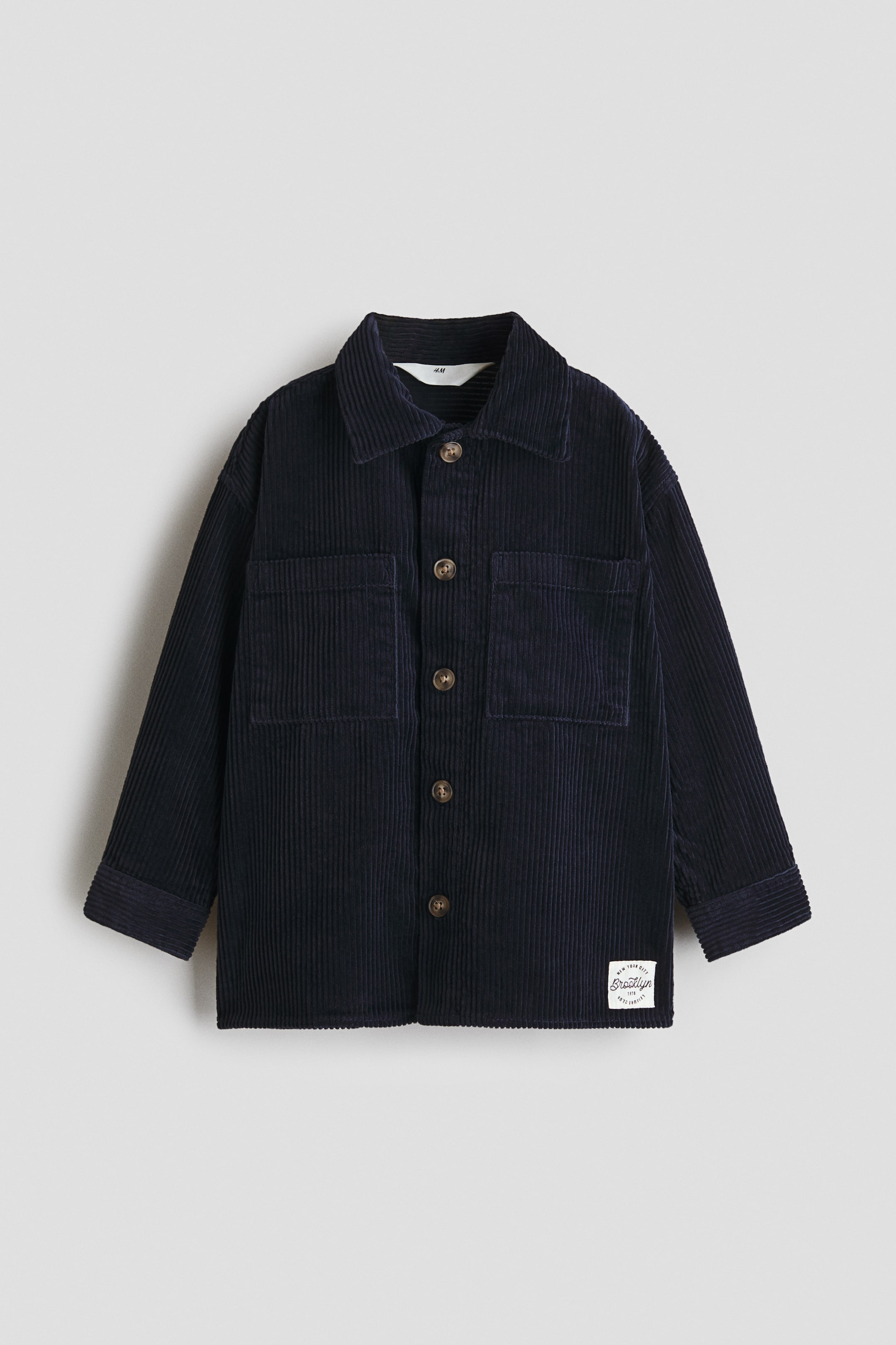 Ver imagen más grande: Una sobrecamisa holgada de pana azul oscuro se muestra de frente, con cuello, botones en la parte delantera y dos bolsillos en el pecho. Está diseñada con hombros caídos, mangas largas y puños abotonados.