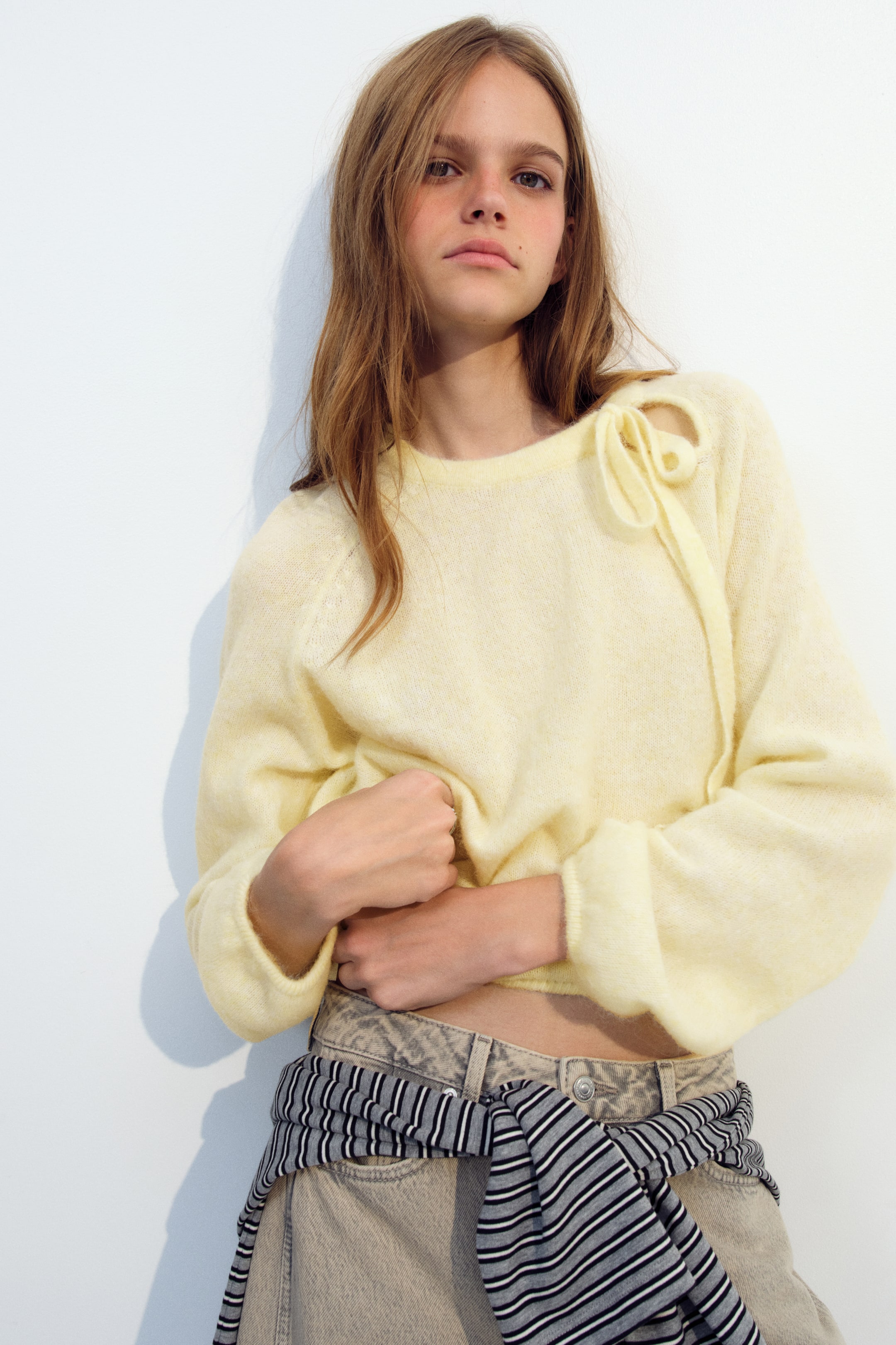 Ampliar la imagen: La modelo lleva un suéter holgado de color amarillo pálido, tejido suave con acabado afelpado, mientras recoge el dobladillo con las manos. El suéter presenta un escote redondo con detalle de moño en el cuello, mangas largas tipo raglán y globo con puños acanalados, y se combina con jeans beige de tiro alto y una camisa a rayas anudada a la cintura.