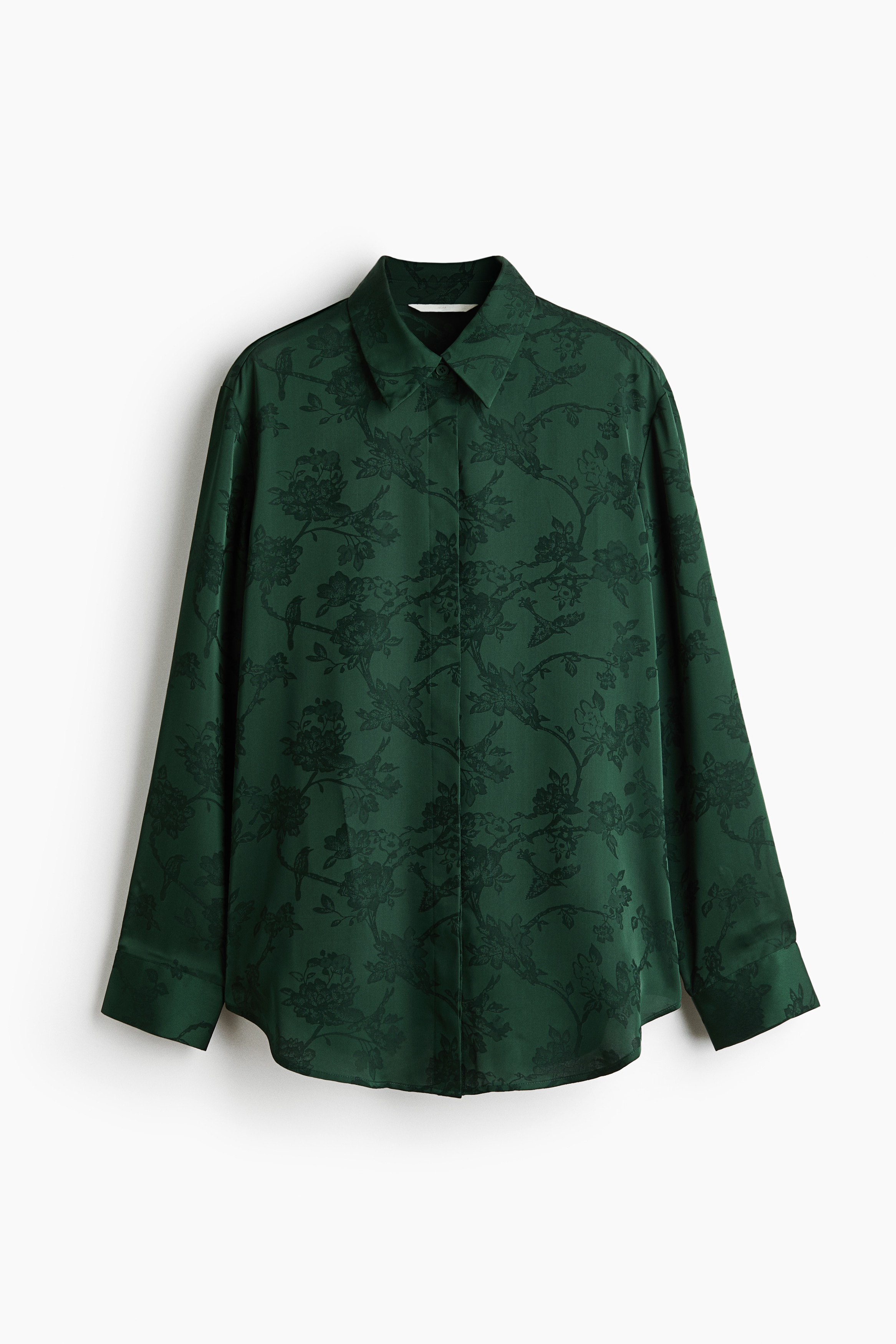 A long shirt in a dark green floral jacquard weave is being worn by the model, featuring a collar, concealed front buttons, and long sleeves with button cuffs. The shirt is styled open at the collar with layered gold necklaces and paired with matching jacquard trousers.