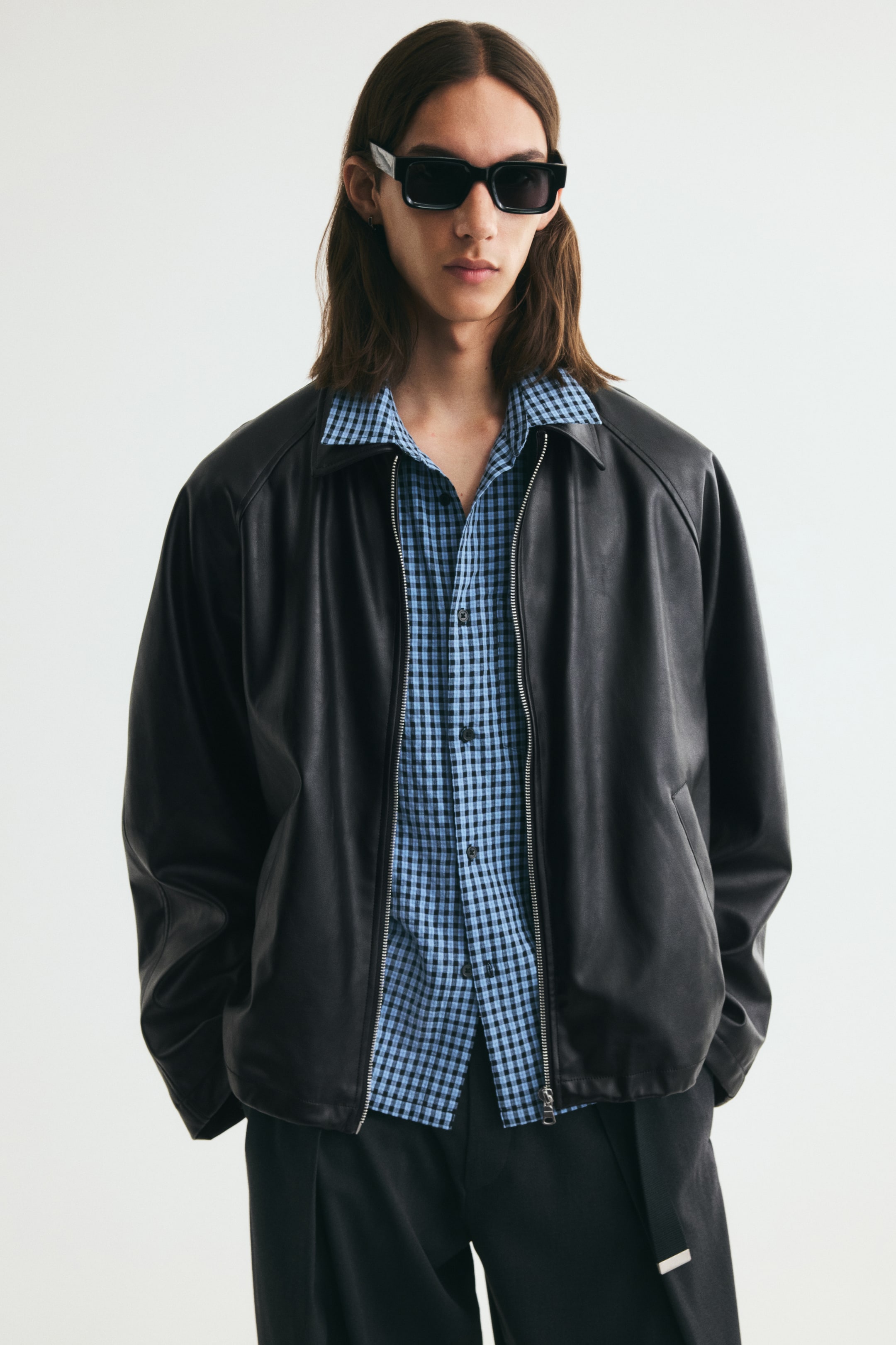 Ver imagen más grande: La modelo lleva una chaqueta negra de tejido recubierto, abierta, sobre una camisa de cuadros vichy azules y negros. La chaqueta presenta un cuello clásico, cierre frontal de cremallera plateada, mangas raglán y bolsillos laterales de ojal.