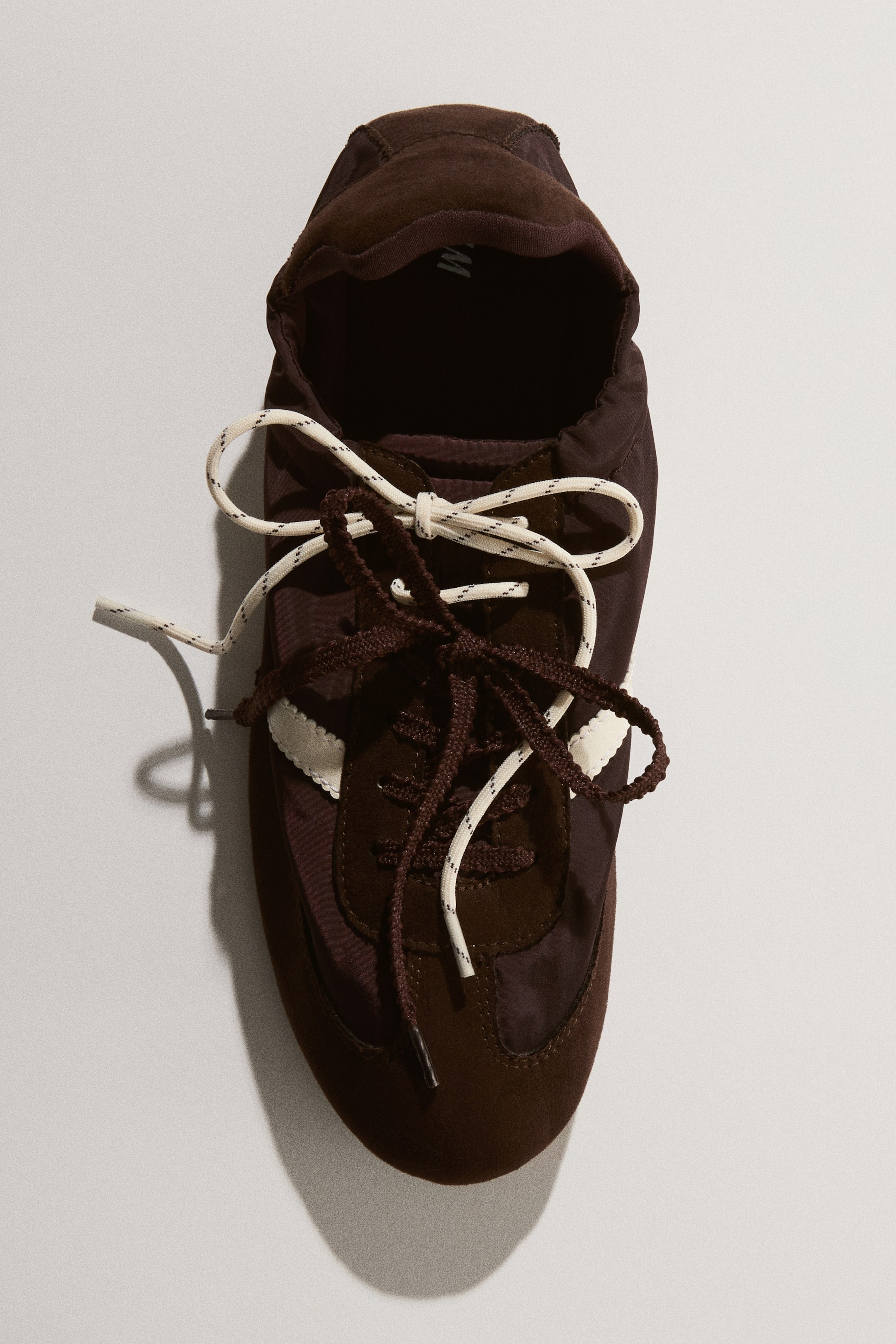 Visualizza immagine più grande: Le sneaker presentano una tomaia marrone scuro, realizzata con pannelli in suede e tessuto. Si distinguono per le strisce ondulate bianche sui lati e per due serie di lacci annodati: una marrone scuro e una bianco sporco con puntinature scure.