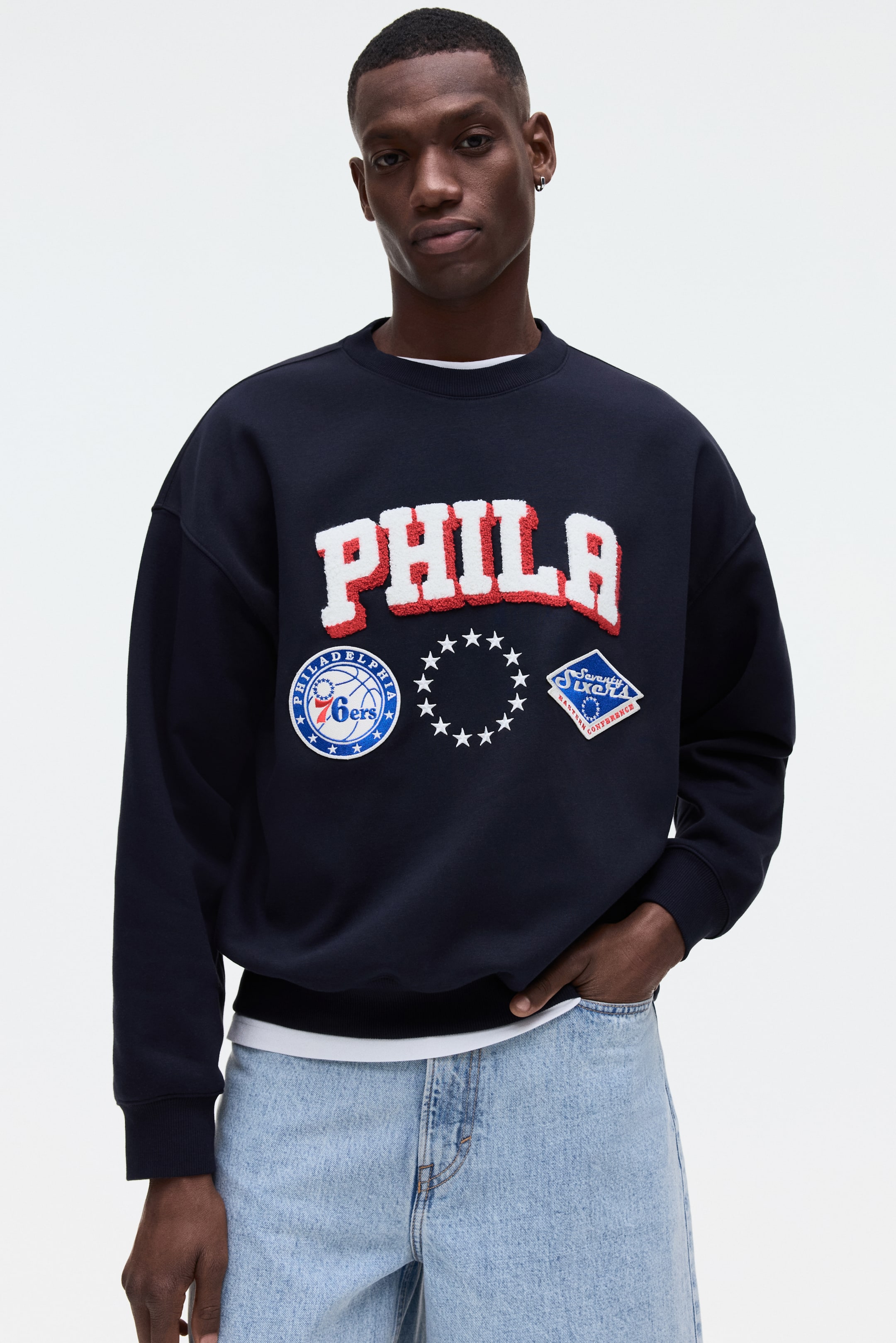 Grösseres Bild ansehen: Ein dunkelblaues Sweatshirt mit Rundhalsausschnitt wird aus einer frontalen Perspektive präsentiert und zeigt ein großes "PHILA"-Applikation in weißem Chenille mit rotem Umriss über der Brust. Begleitet wird es von einem runden Philadelphia 76ers Logo-Patch, einem Kreis aus weißen Sternen-Patch und einem rautenförmigen Seventy Sixers-Patch darunter. Das Sweatshirt ist mit gerippten Bündchen und einem gerippten Saum gestaltet und wird über einem weißen T-Shirt getragen, das am Ausschnitt und am Saum sichtbar ist.