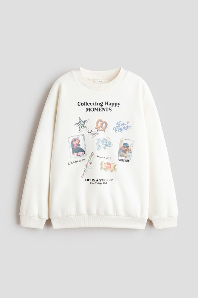Una felpa morbida color bianco sporco Ã¨ mostrata frontalmente, con scollo rotondo rifinito a coste, spalle scese e maniche lunghe con polsini e orlo a coste. Sul davanti Ã¨ presente una stampa colorata con la scritta "Collecting Happy MOMENTS" circondata da varie grafiche in stile sticker, tra cui ritratti di cani, una stella e un pretzel. - 1