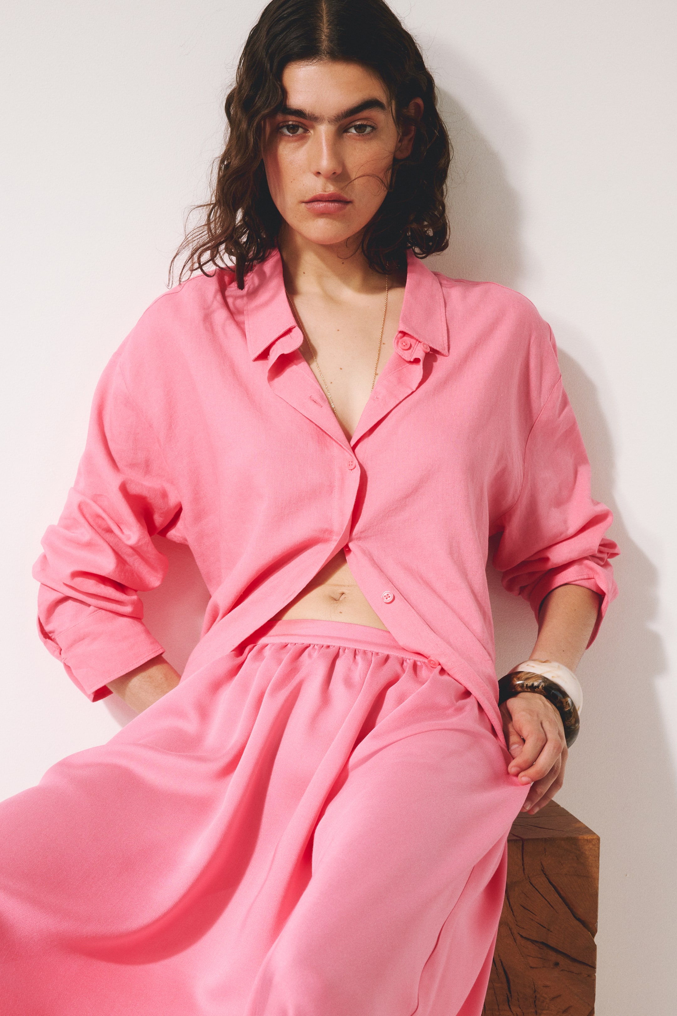 View larger image: A pink button-up shirt and matching wide-leg pants are displayed from a slightly angled front view. The shirt features a pointed collar and long sleeves rolled up, with several buttons undone at the front, while the pants highlight a gathered waist and a loose, flowing drape.
