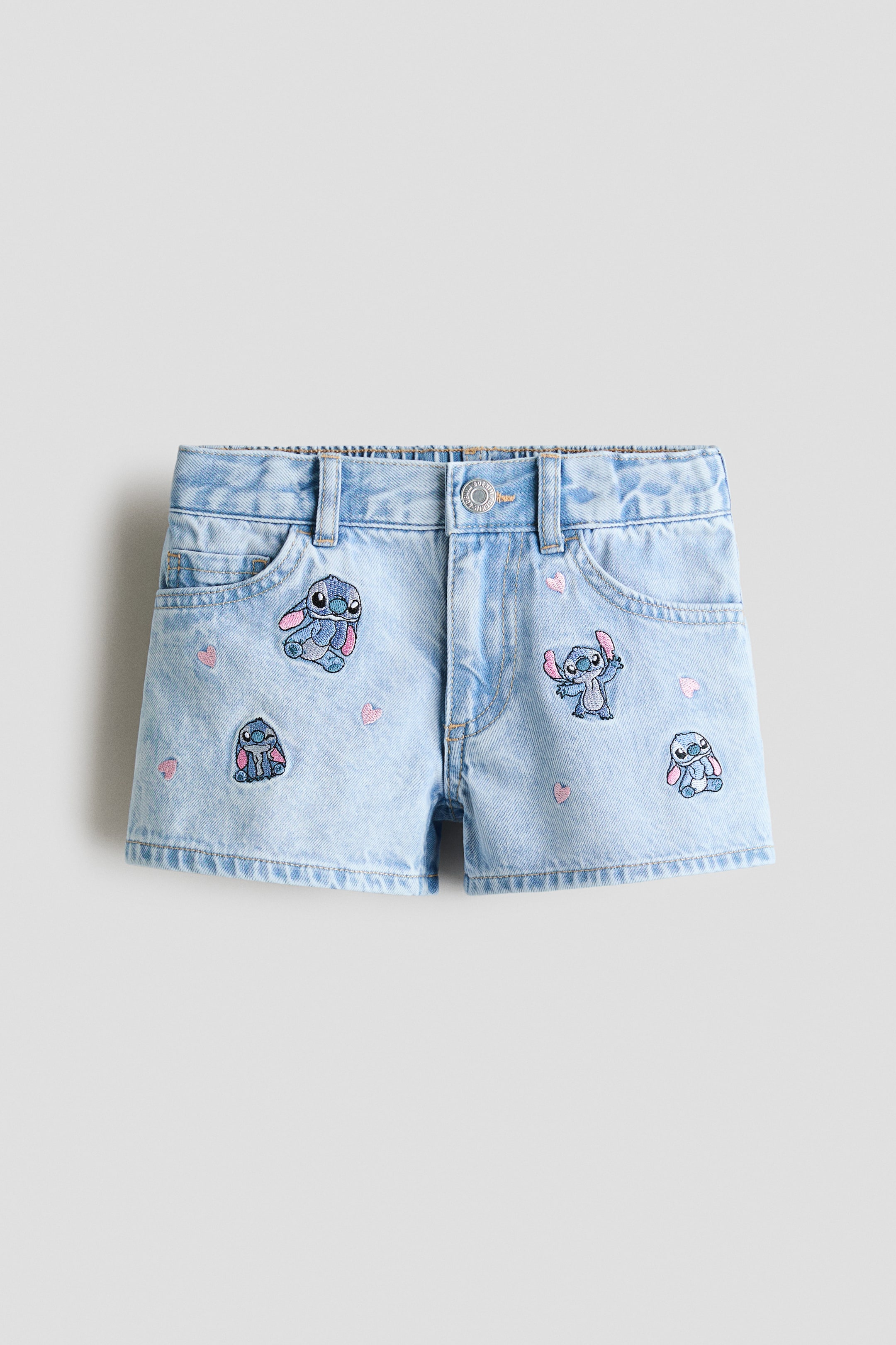 View larger image: Light blue cotton denim 5-pocket shorts are shown from the front, featuring an adjustable, elasticated waist with belt loops and a zip fly with a press-stud. The shorts are adorned with multiple printed motifs of a blue character and small pink hearts, while displaying two front pockets and a coin pocket.