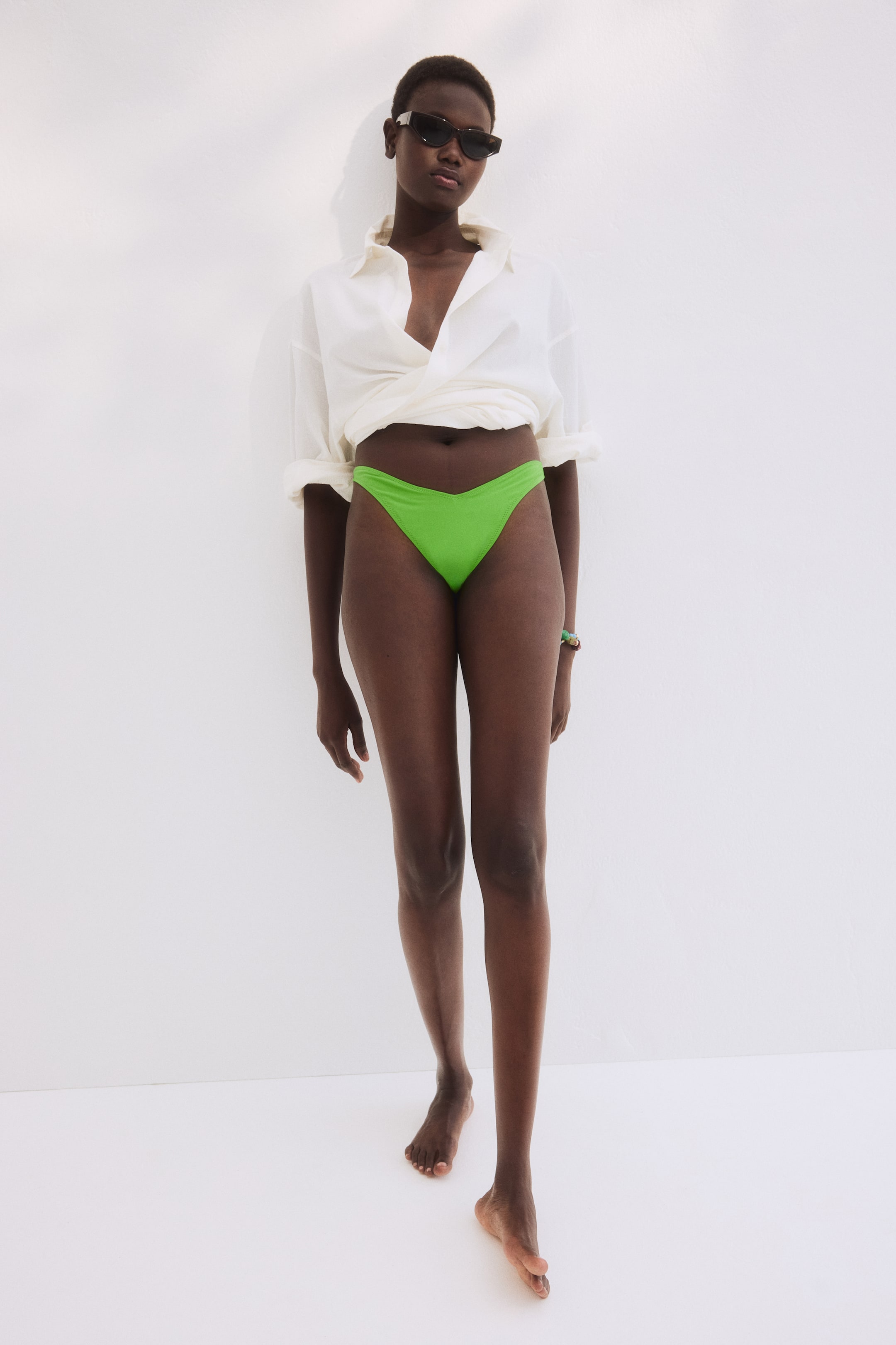 View larger image: The model is showcasing vibrant green thong bikini bottoms with a low, V-shaped waistline. A white button-down shirt is styled loosely over the bikini bottoms.