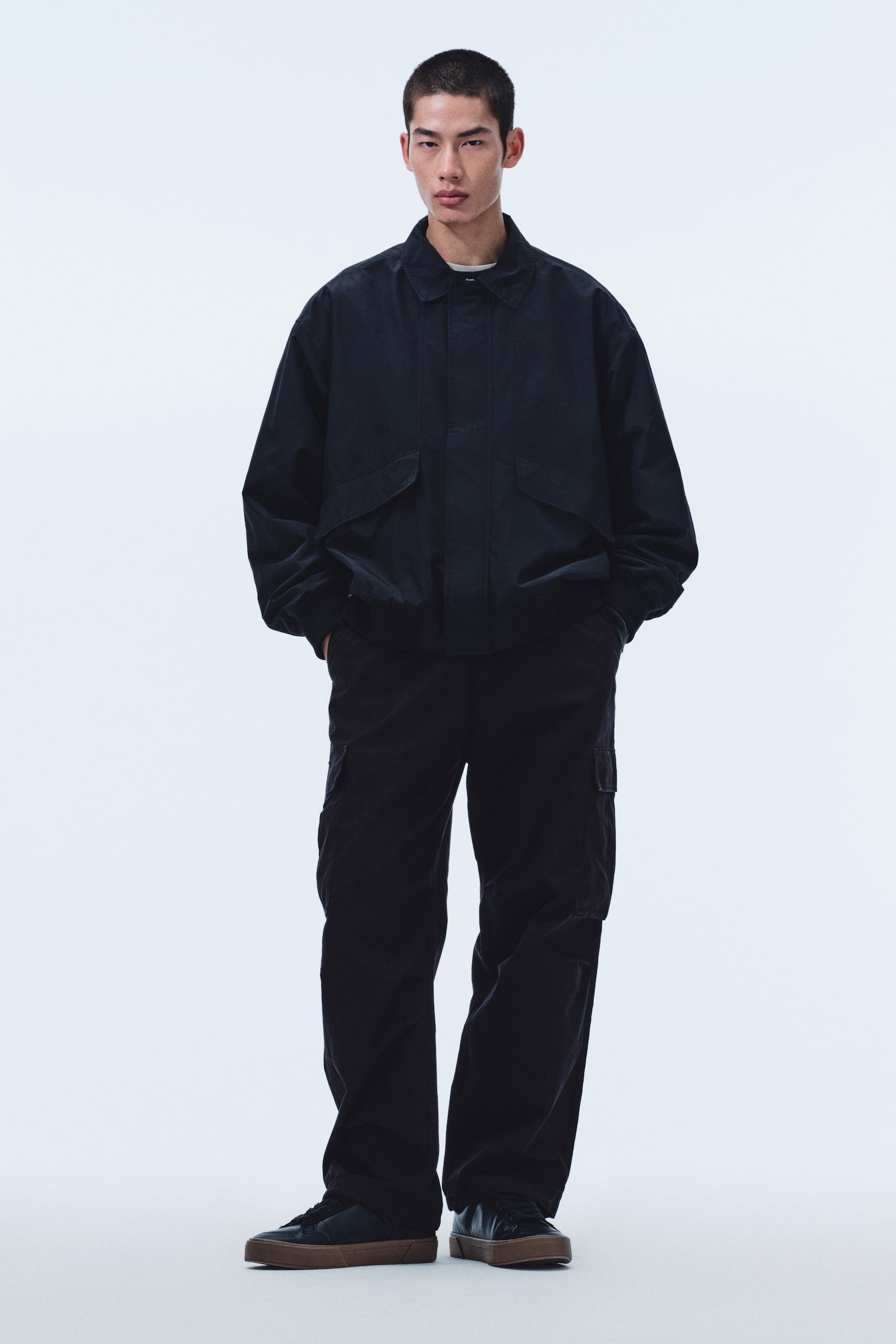 A dark, loose-fitting jacket features a pointed collar and two large flap pockets on the front, with a light-coloured undershirt visible at the neckline. Dark, loose-fitting cargo trousers with side pockets are paired with dark shoes with thick brown soles. The entire ensemble is presented in a front-facing view. - 1