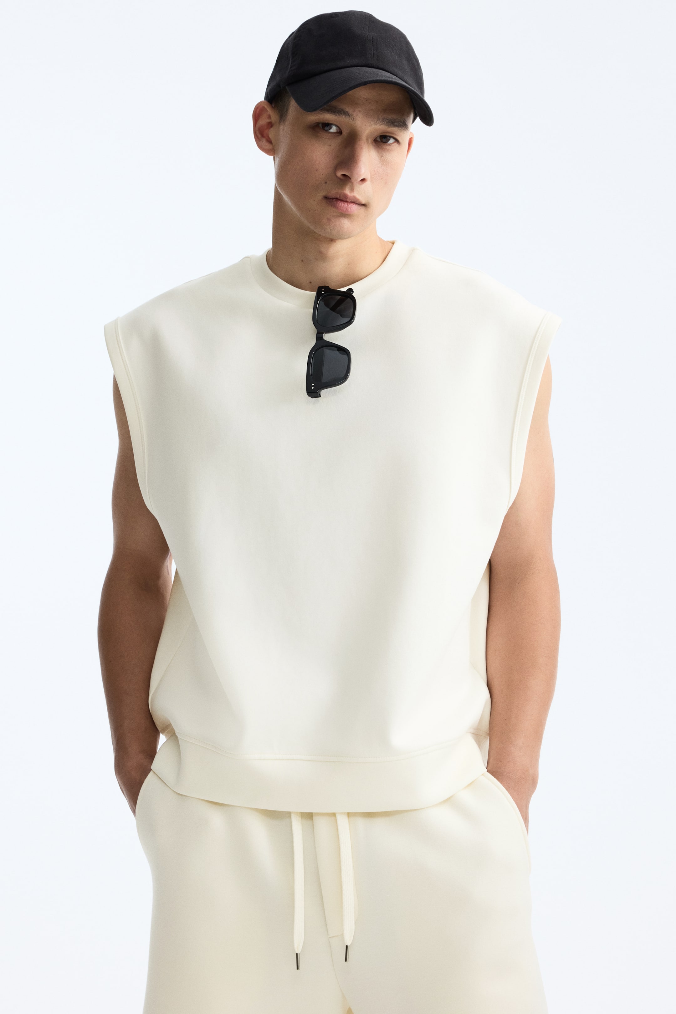 View larger image: A cream-coloured sleeveless top and matching sweatpants are being presented as a set. The top is designed with a crew neck, wide armholes, and a ribbed hem, while black sunglasses are resting on its neckline. The sweatpants are designed with a visible drawstring waistband.