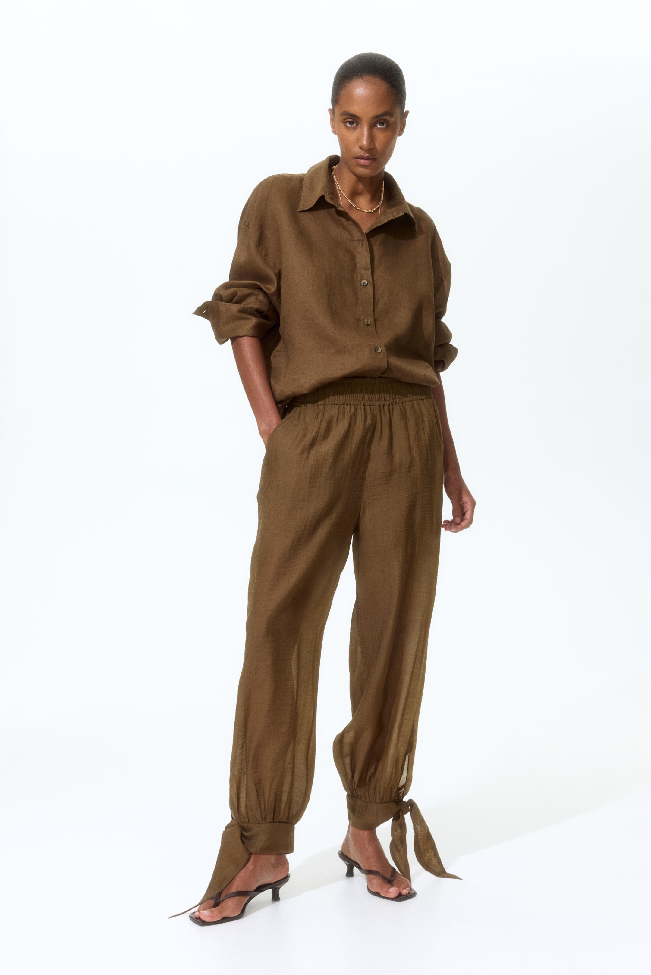 View larger image: Presenting an earthy brown linen shirt paired with matching pants. The shirt, designed for a relaxed fit, showcases a pointed collar, button-down front, and rolled long sleeves, partially tucked into the pants. The pants feature an elasticated waistband, side pockets, and gathered ankles with ties that wrap around the feet.