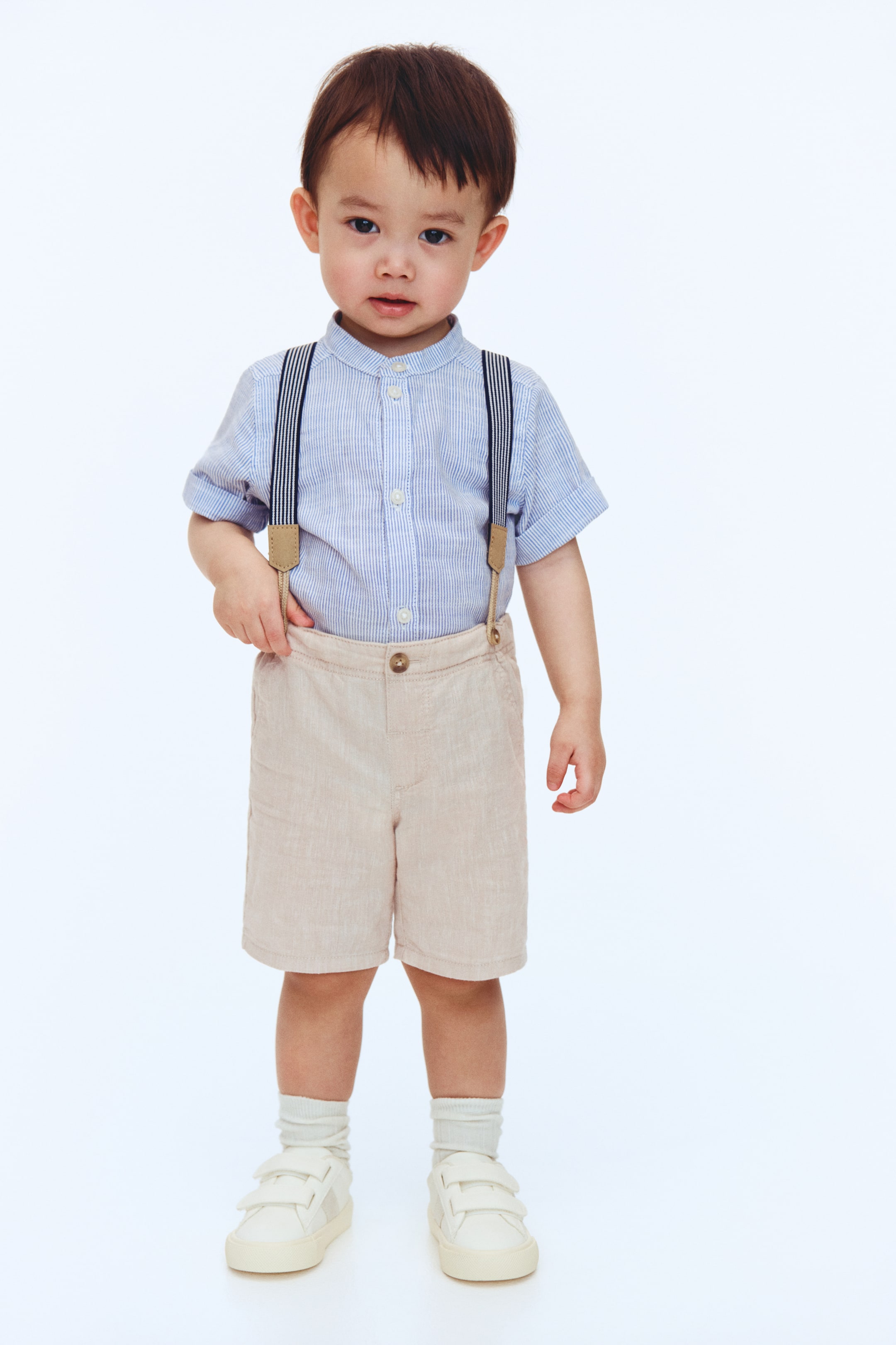 Ver imagen más grande: Un conjunto infantil presenta una camisa de rayas azul claro y blanco de manga corta con cuello mao, combinada con tirantes de rayas azul oscuro y blanco que se sujetan a unos pantalones cortos texturizados de color beige claro. Calcetines de canalé blancos y zapatillas crema con cierre de velcro completan el look, todos orientados hacia el frente.