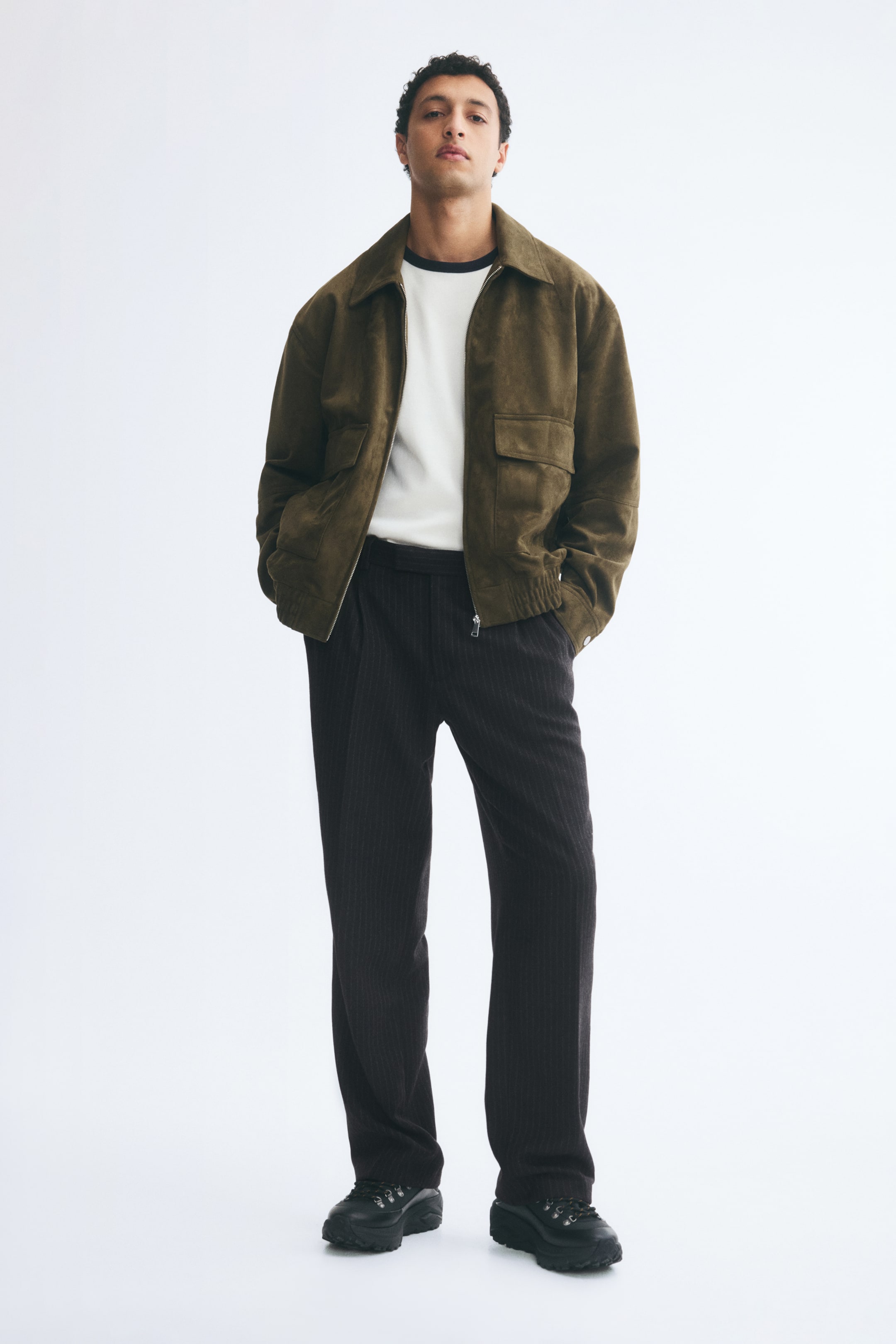 Voir une image plus grande: Le mannequin porte un pantalon sur mesure en flanelle à fines rayures foncées, avec une coupe décontractée, des jambes droites avec des plis marqués à l’avant et de subtils plis à la taille. Le pantalon est agencé avec un t-shirt blanc, un blouson zippé vert olive et des chaussures noires.