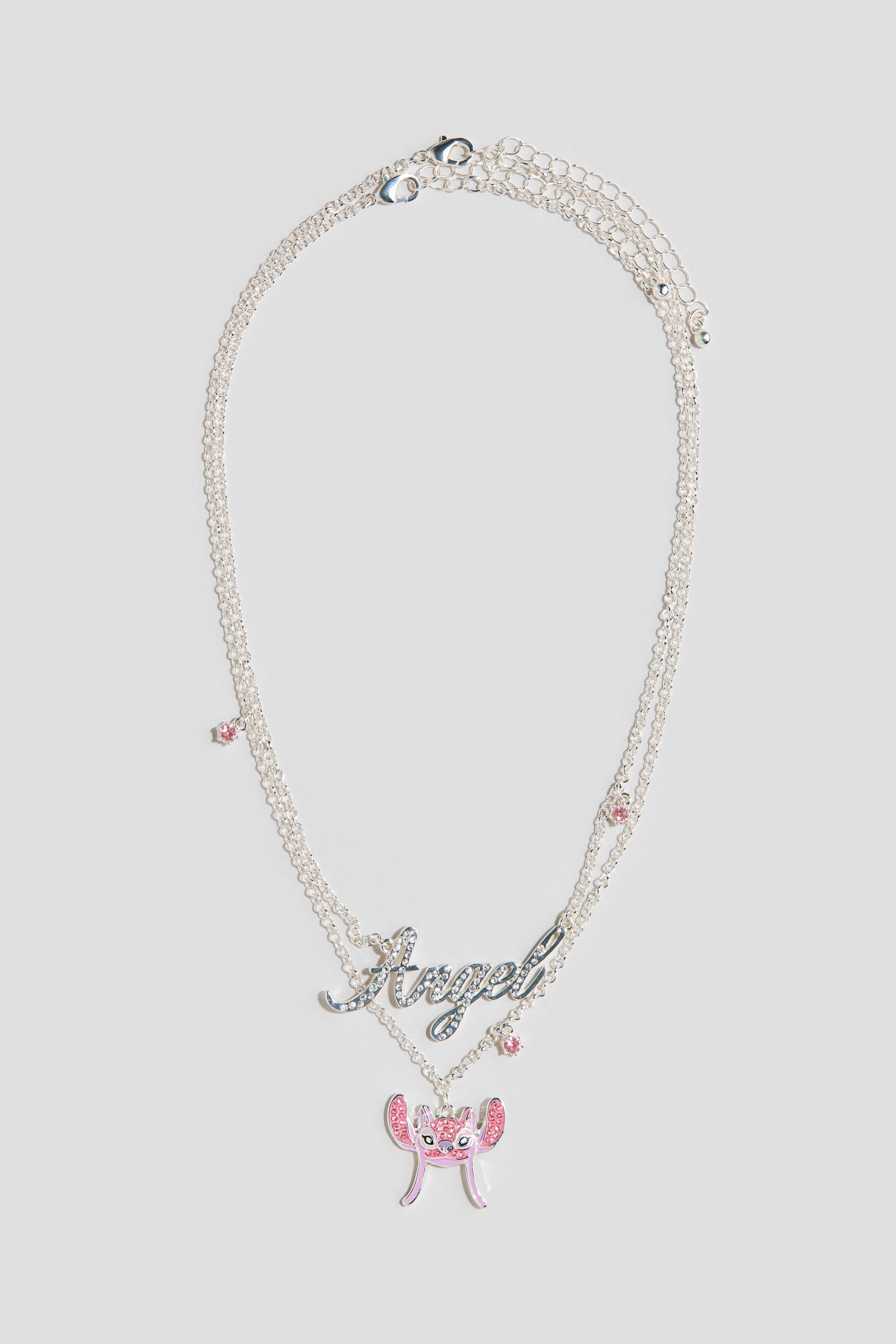 Visa större bild: Ett lager-på-lager halsband i silverton visas framifrån, med två kedjor, båda prydda med små rosa stenar. Den övre kedjan visar upp ett 'Angel'-hänge i skrivstil dekorerat med klara stenar, medan den nedre kedjan har ett rosa och silvertonat karaktärshänge utsmyckat med rosa stenar.