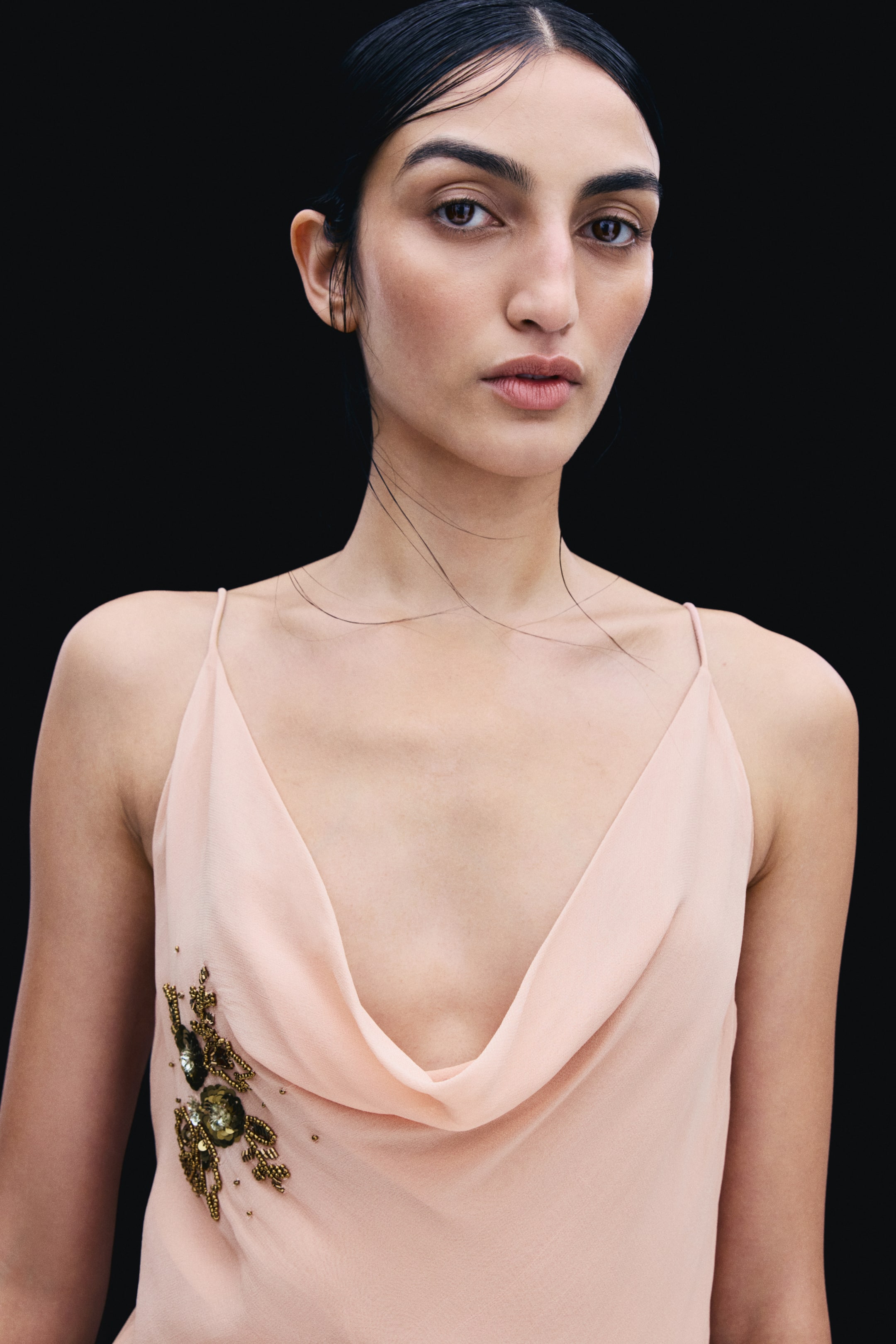 View larger image: The model is wearing a peach-coloured chiffon dress featuring a draped cowl neckline, thin spaghetti straps, and a gold-toned sequin and bead floral embellishment.