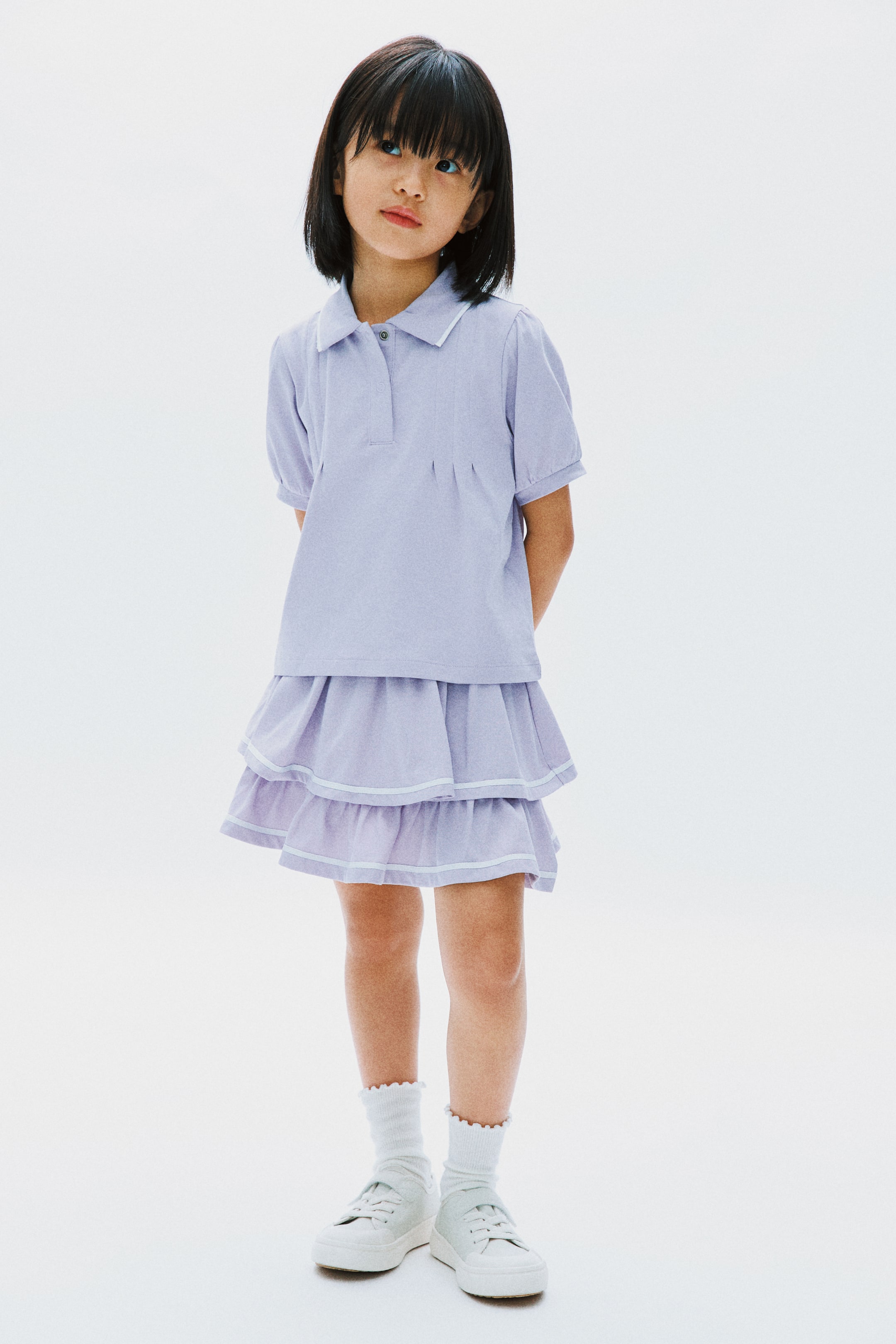 View larger image: Model memakai baju polo berwarna lavender dengan lengan pendek, menampilkan kolar bertrim putih dan plaket dua butang, sebagai sebahagian daripada set yang dipadankan dengan skirt bertingkat yang sepadan dengan trim putih pada setiap tingkat, digayakan bersama stokin buku lali beropol putih dan kasut sneakers bertali putih.