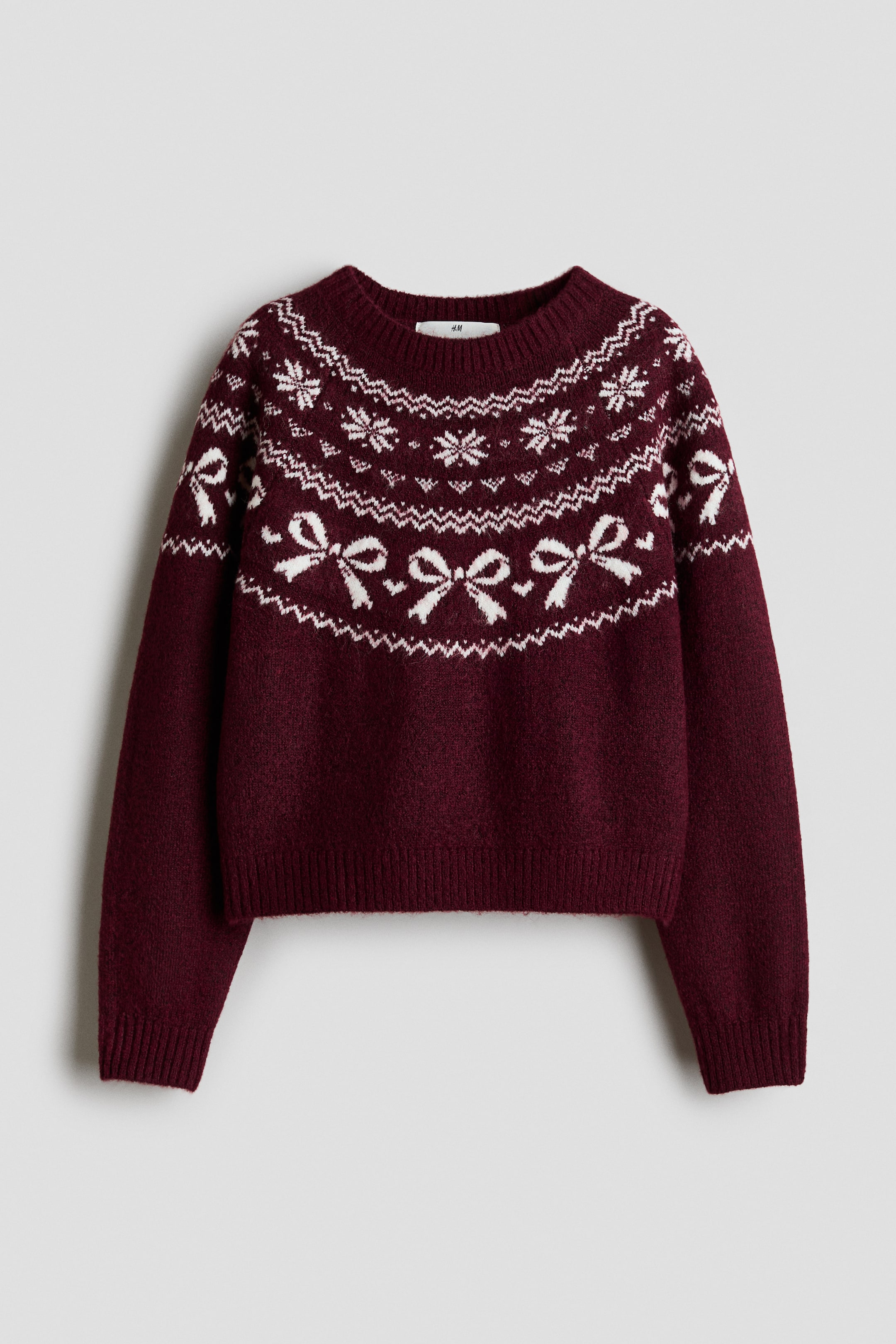 Voir une image plus grande: Un chandail en tricot bordeaux est présenté de face, avec un motif Fair Isle blanc de flocons de neige et de nœuds sur l’empiècement et le haut des manches. Le chandail est doté d’un col rond côtelé, de manches longues avec poignets côtelés et d’un ourlet côtelé.