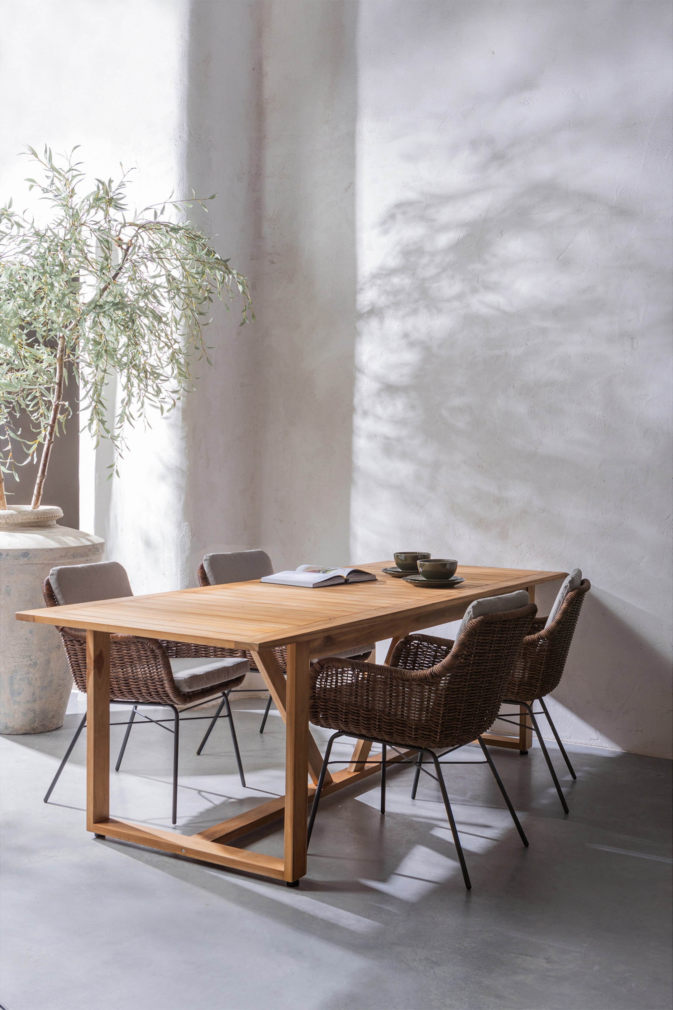 Agrandir l'image: Une table à manger rectangulaire en bois, avec un dessus à lattes et une base présentant des supports inclinés, est présentée sous un angle avant-droit, avec un livre ouvert et deux bols de service sombres avec des assiettes sur sa surface. Quatre chaises, construites avec des sièges et dossiers en osier brun, des coussins gris clair et des pieds en métal foncé, sont disposées autour de la table.