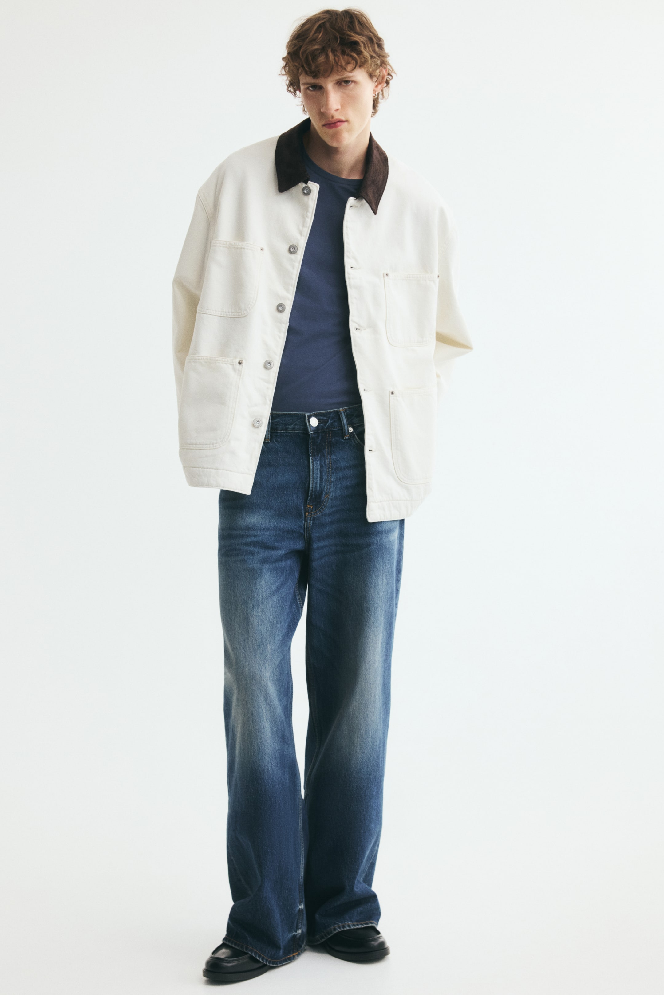 View larger image: An off-white denim chore jacket featuring a contrasting dark brown corduroy collar is worn open by the model. The jacket has patch pockets on the chest and lower front, and is layered over a dark blue T-shirt and blue jeans.