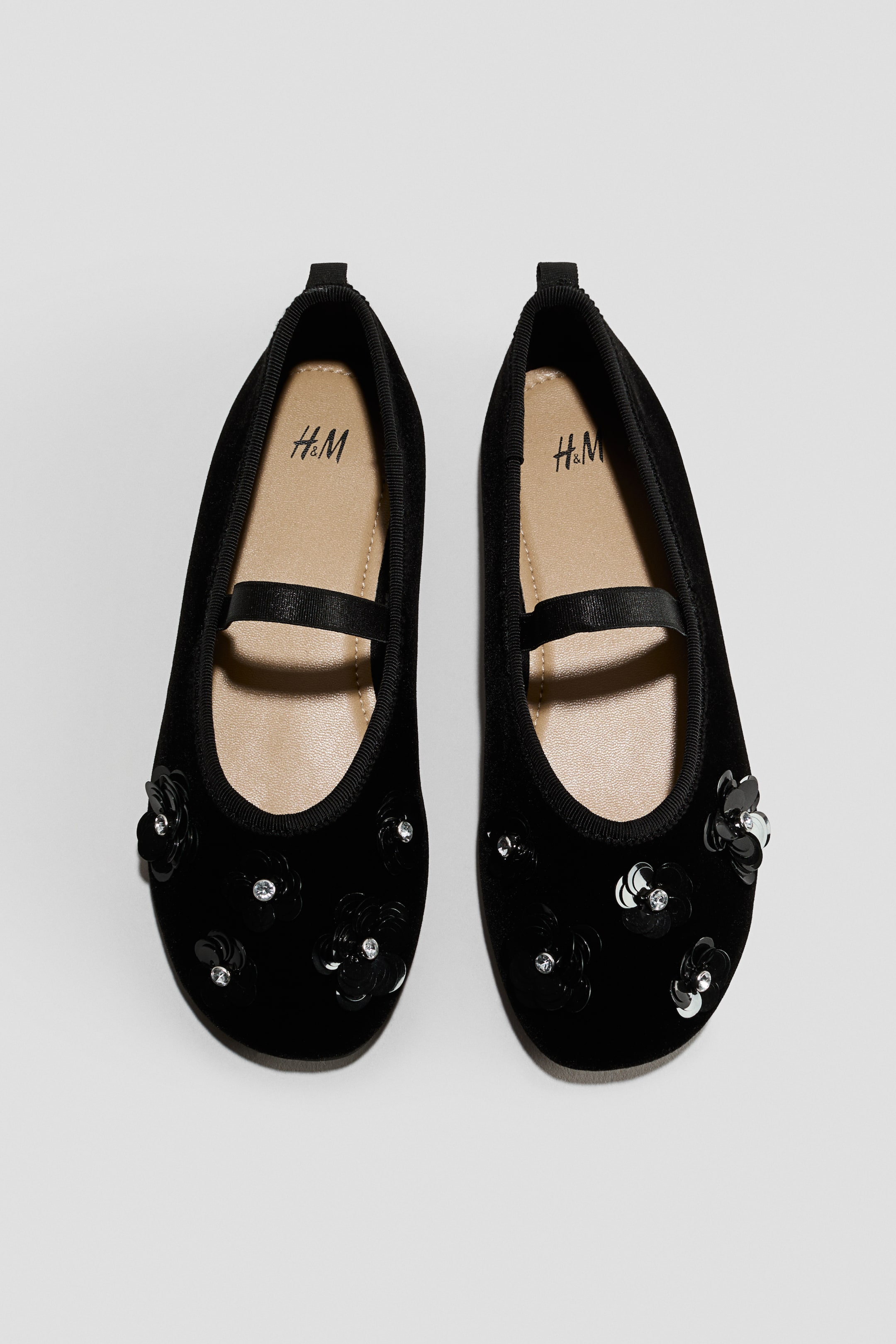 View larger image: A pair of black ballet pumps is shown from a top-down angle, featuring flower-shaped embellishments made of black sequins and clear rhinestones on the toes. The shoes are designed with a black elasticated foot strap across the instep, a matching grosgrain trim around the opening, a small loop at the heel, and light beige insoles.