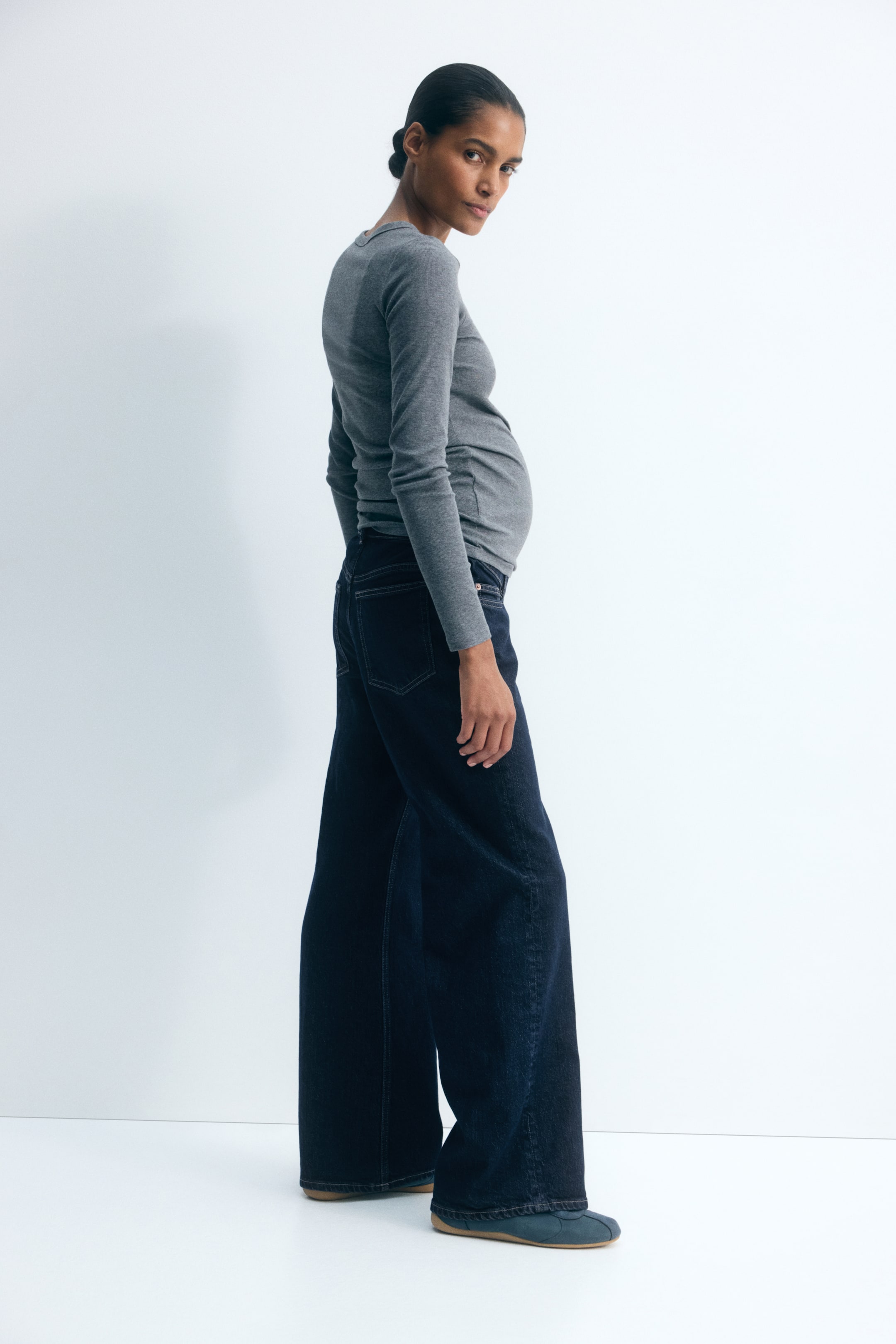 View larger image: The model is wearing dark wash maternity jeans with a relaxed fit and wide straight legs. The jeans are styled with a fitted, long-sleeved gray top layered over the waistband.