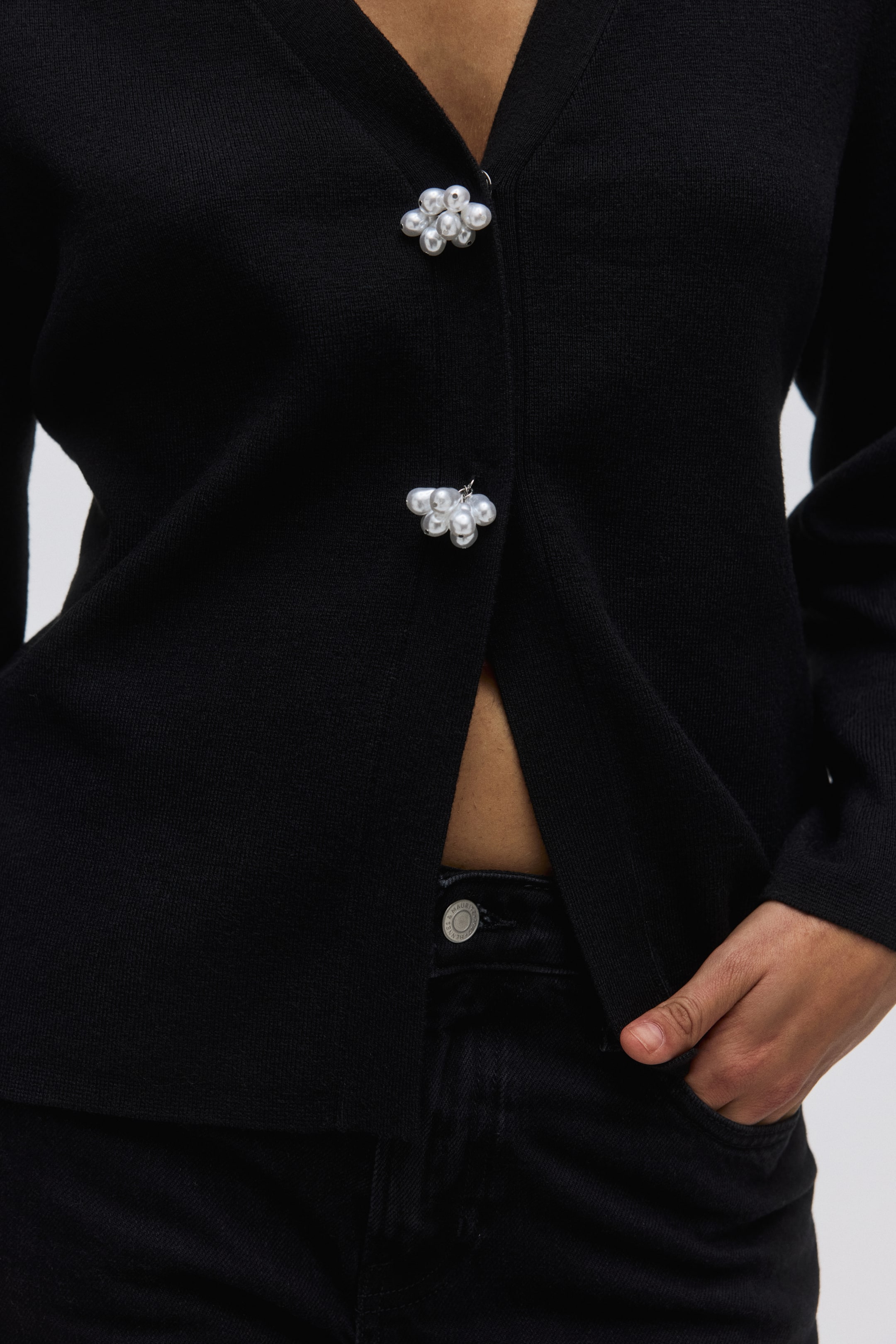 View larger image: A black knit cardigan is presented from the front, featuring a V-neck and long sleeves. It fastens at the front with two decorative pearl cluster buttons and is styled open, revealing the midriff.