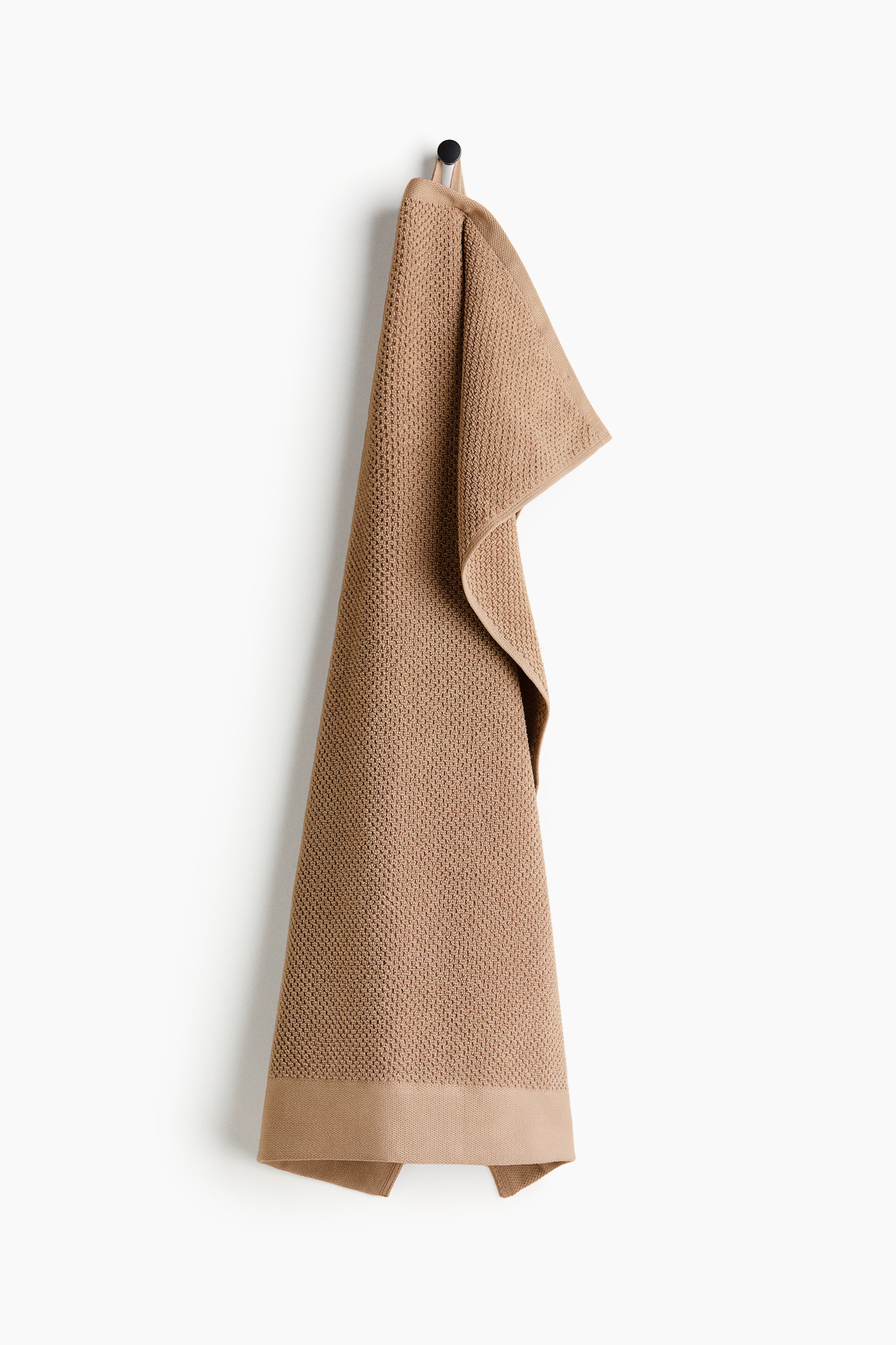 View larger image: A light brown hand towel is hung vertically to display its front; it features a textured waffle weave with a smooth, flat band along the bottom edge. A loop at the top secures the towel to a hook.
