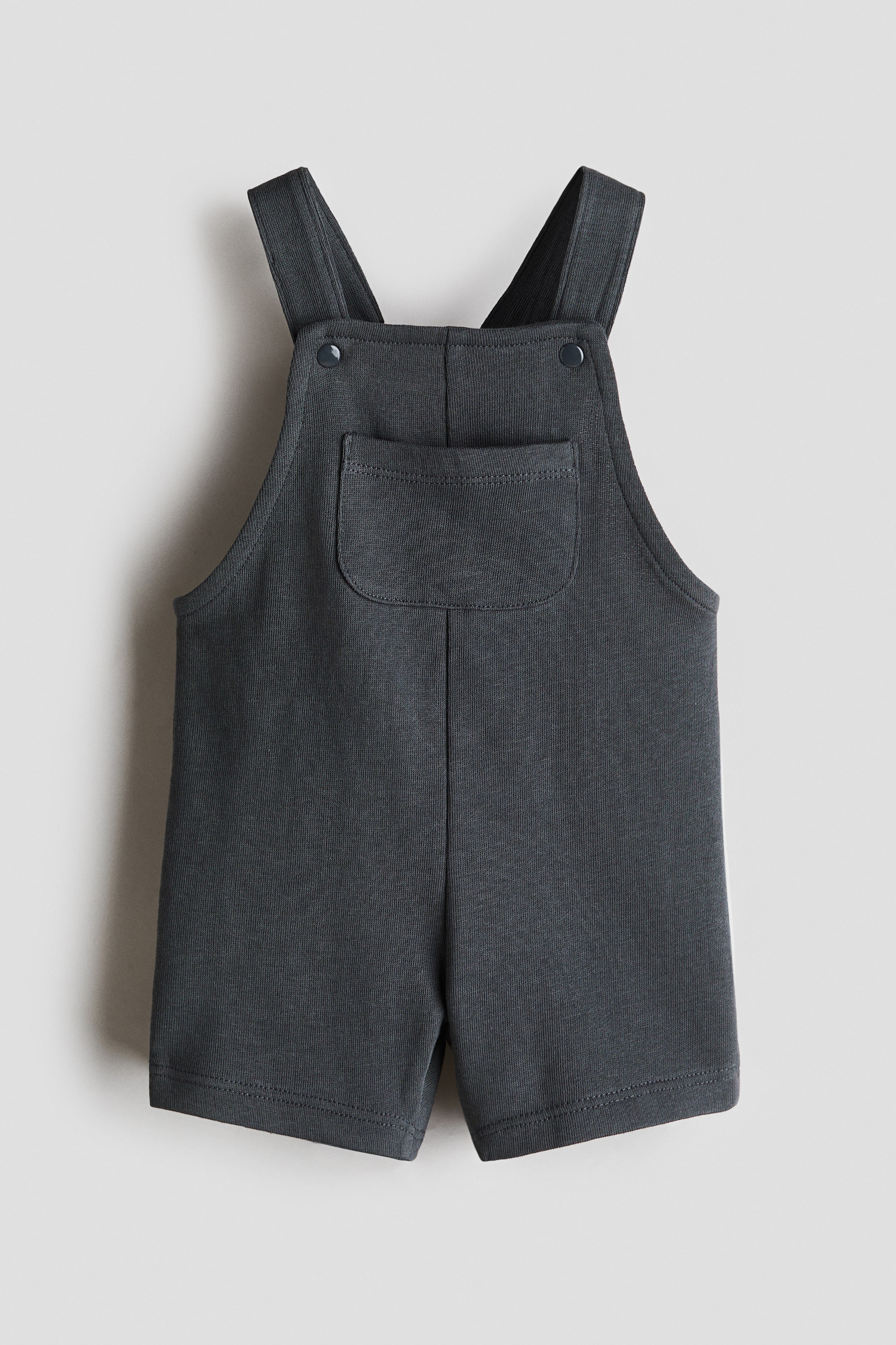 View larger image: Dungaree shorts are depicted from the front, made from a soft, lightweight sweatshirt fabric. They showcase shoulder straps with press-stud fastening, a rounded patch chest pocket on the bib, and short legs.