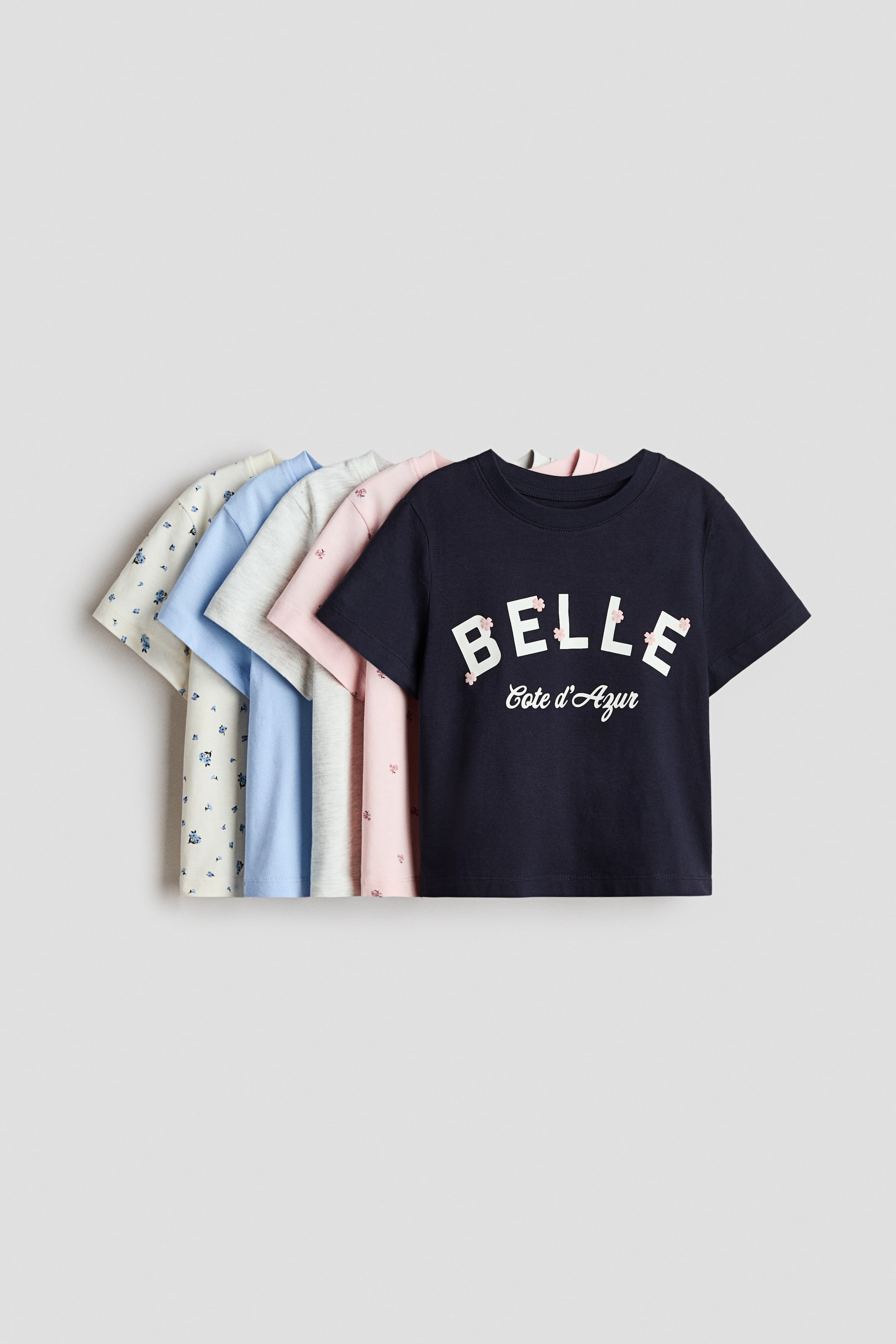 Agrandir l'image: Cinq t-shirts à manches courtes sont disposés, montrant leurs devants et présentant des bordures côtelées autour des encolures. Le t-shirt le plus à l'avant est bleu foncé avec le texte "BELLE Cote d'Azur" et des détails floraux roses, tandis que les t-shirts partiellement visibles derrière sont en rose clair, gris clair, bleu clair et crème, certains avec de petits motifs floraux.