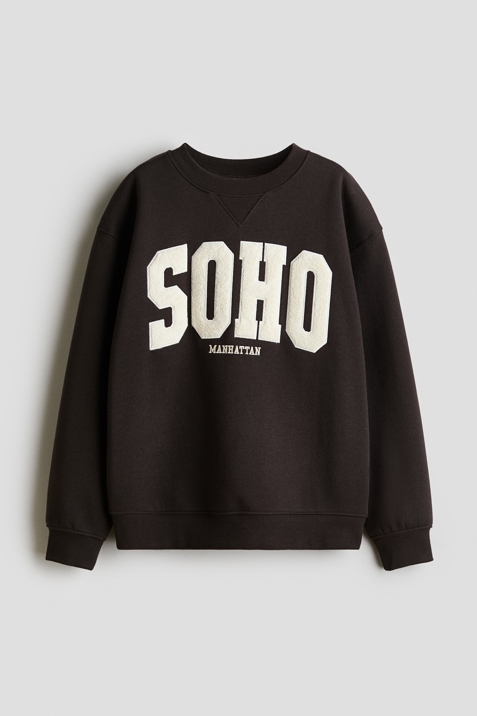 Una felpa girocollo marrone scuro Ã¨ mostrata frontalmente, con colletto a coste con dettaglio a V, polsini a coste e orlo a coste. Sul petto sono presenti grandi lettere applicate testurizzate color crema che compongono la scritta "SOHO", con la parola "MANHATTAN" ricamata in caratteri piÃ¹ piccoli subito sotto.