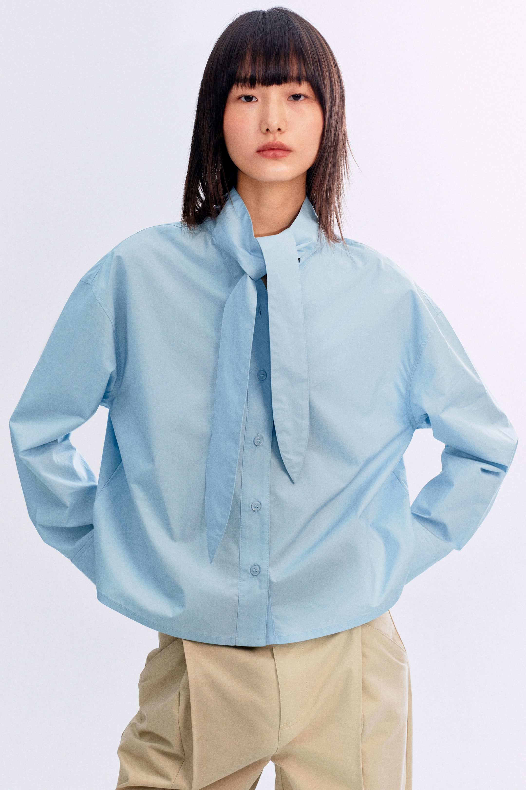 이미지 확대 보기: 연한 파란색 버튼다운 셔츠는 앞쪽에 매듭지어진 긴 타이가 있는 높은 칼라가 특징입니다. 긴 소매와 단추가 달린 커프스, 여유 있는 핏으로 디자인되어 있으며, 연한 갈색 바지 바로 위에 걸쳐져 있습니다.